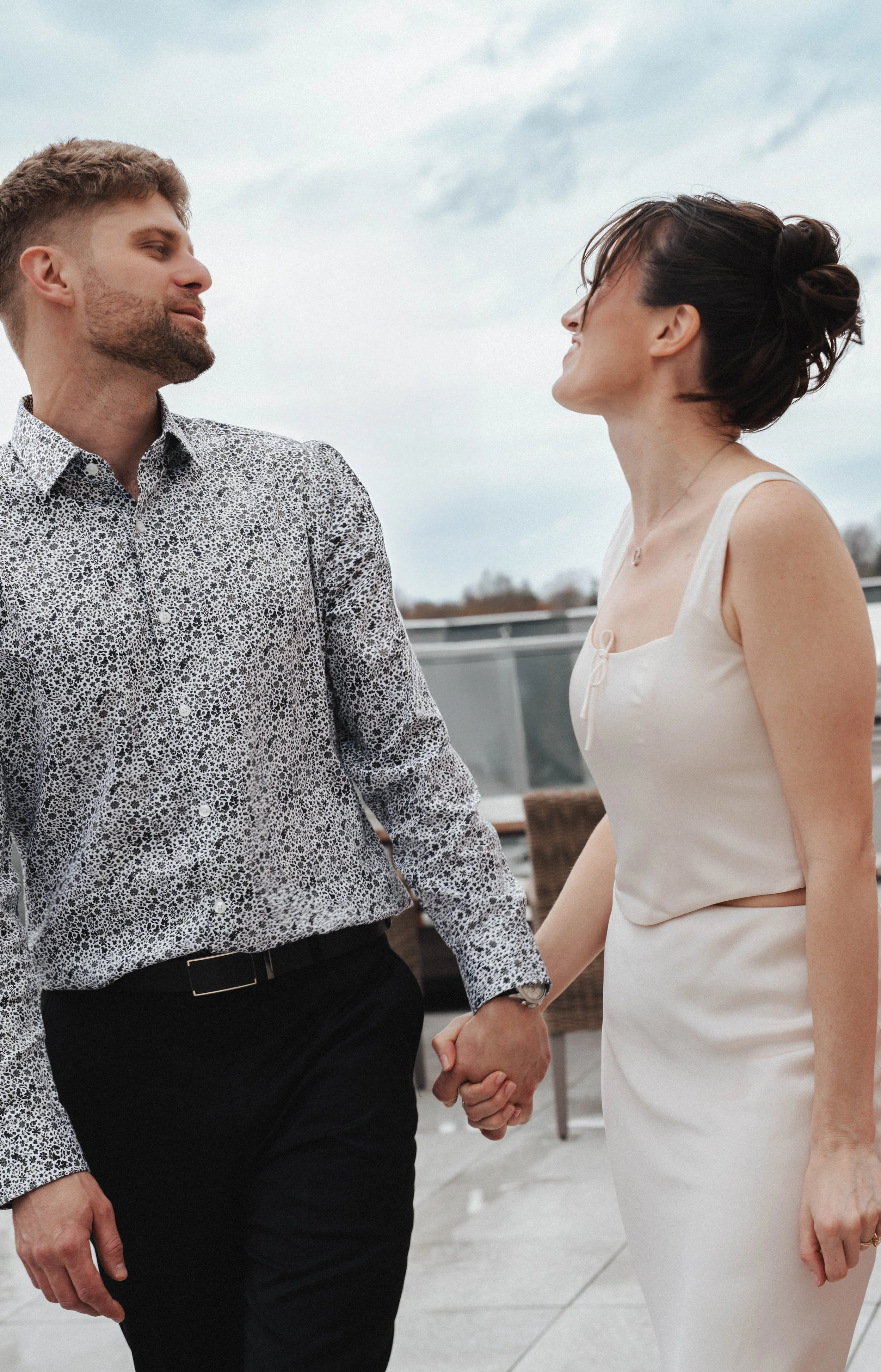 A couple is holding hands and looking at each other, outdoors on a cloudy day, dressed in semi-formal attire.