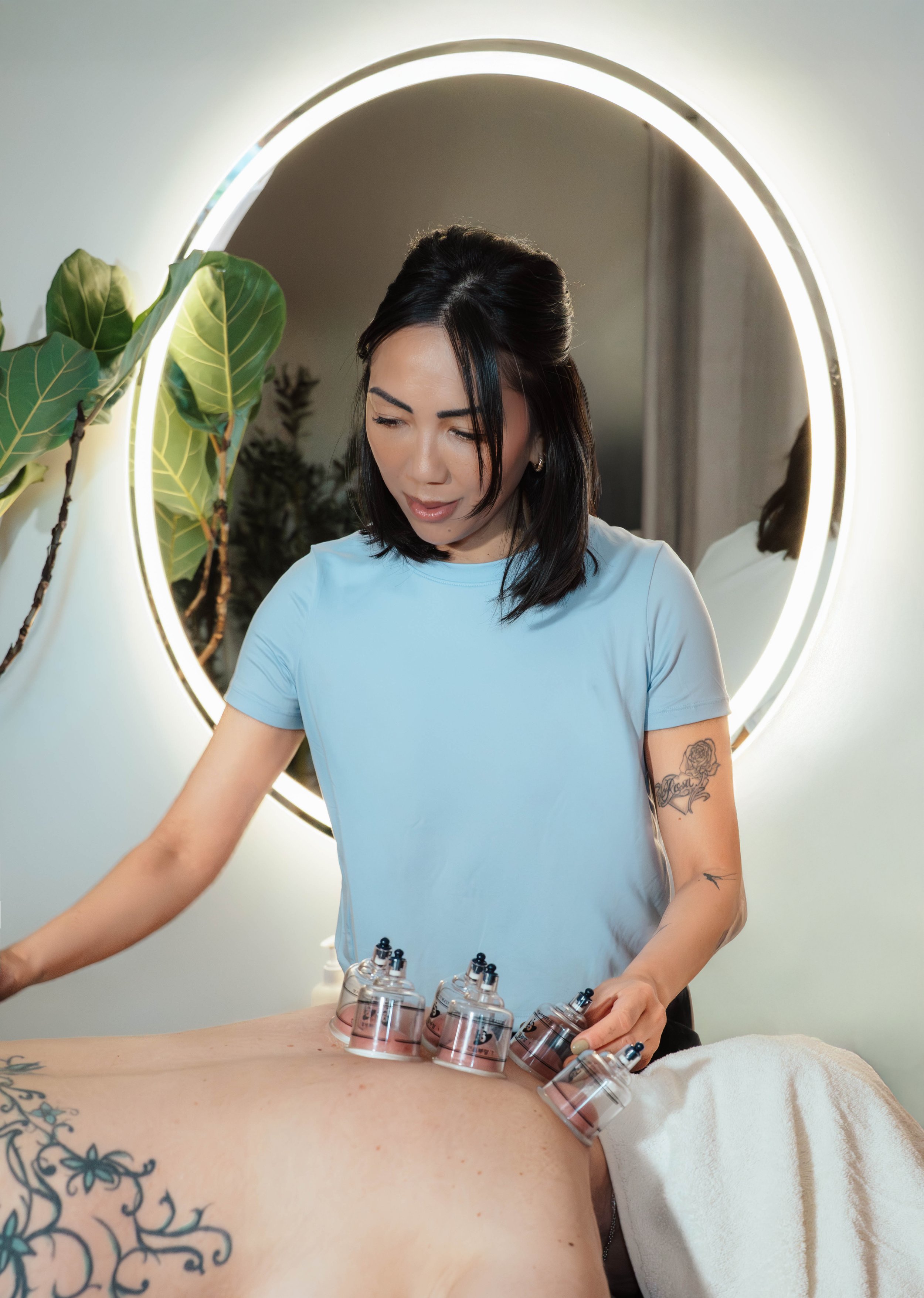 A woman in a light blue shirt performs a cupping therapy session on a patient's back in a clinic with a round illuminated mirror and green plant in the background.