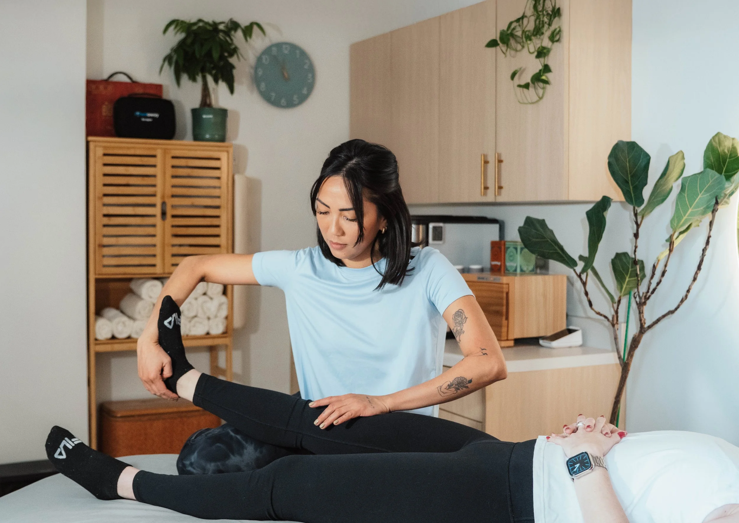 A woman giving a shoulder and arm massage to a person lying on a massage table in a room with wooden cabinets and plants.