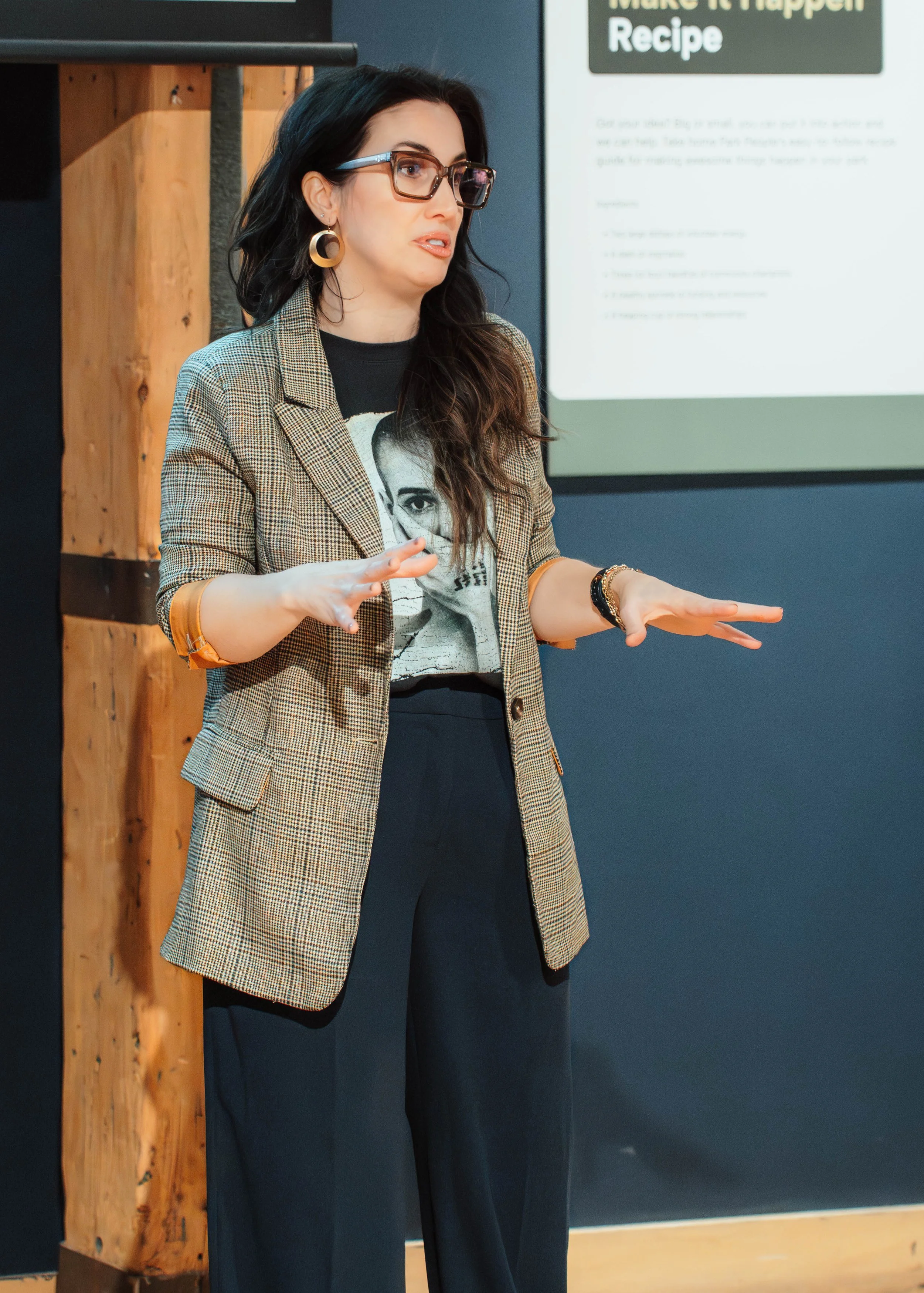 A woman with long dark hair, wearing glasses, a plaid blazer, a graphic T-shirt, and black pants, is giving a presentation in front of a screen with a slide titled 'Make It Happen Recipe'.