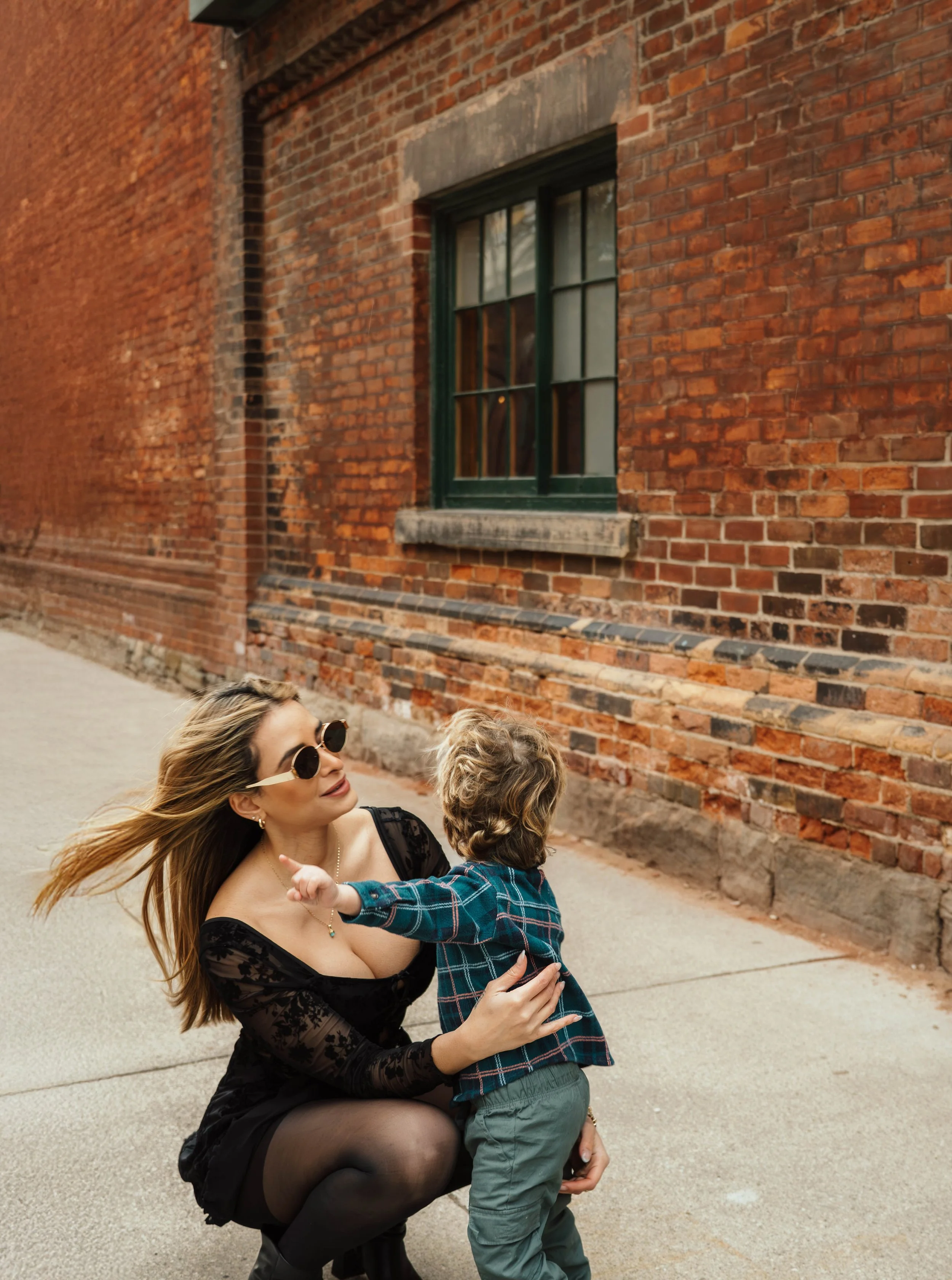 A woman with sunglasses kneeling and holding a young boy with curly hair in a plaid shirt on a sidewalk beside a red brick wall with a window.