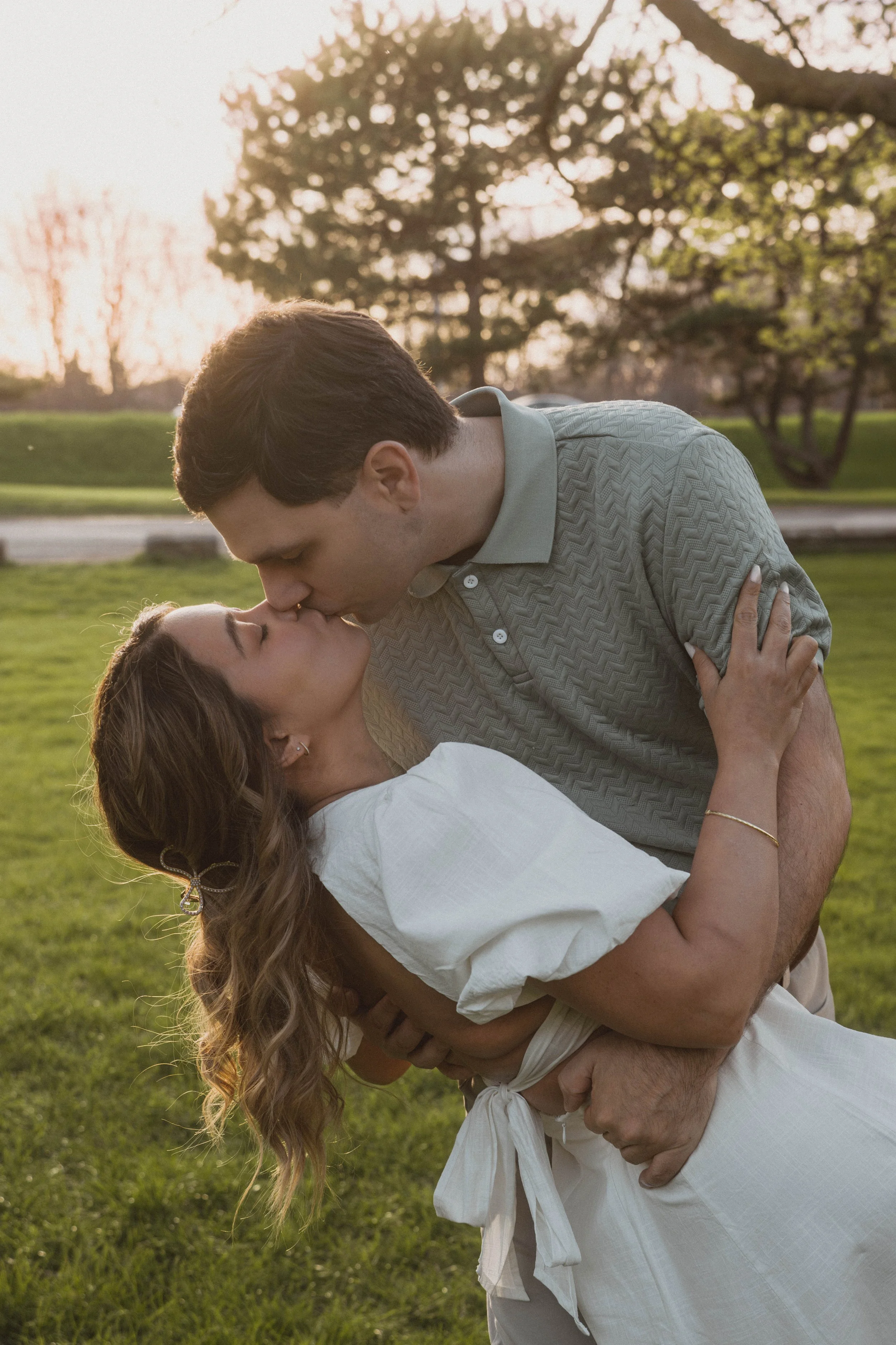 A couple dancing outdoors in a park during sunset, with the man dipping the woman and about to kiss, surrounded by green grass and trees.