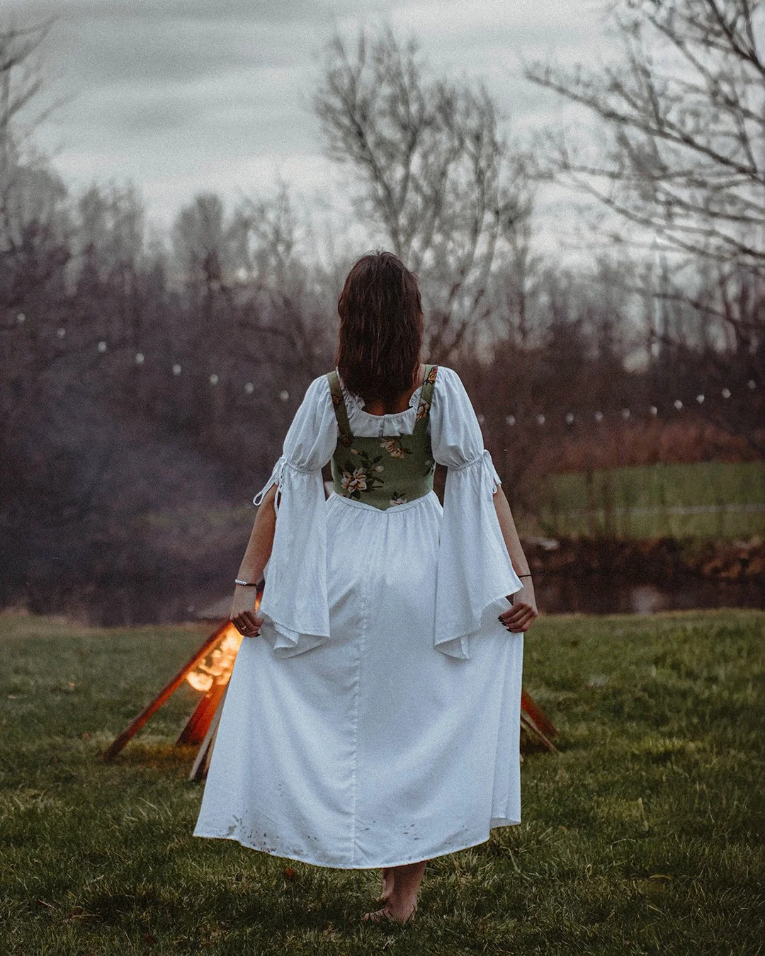 A woman dressed in a white skirt, white blouse, and green floral top standing on grass, facing away, holding the edges of her skirt, with a campfire burning behind her.