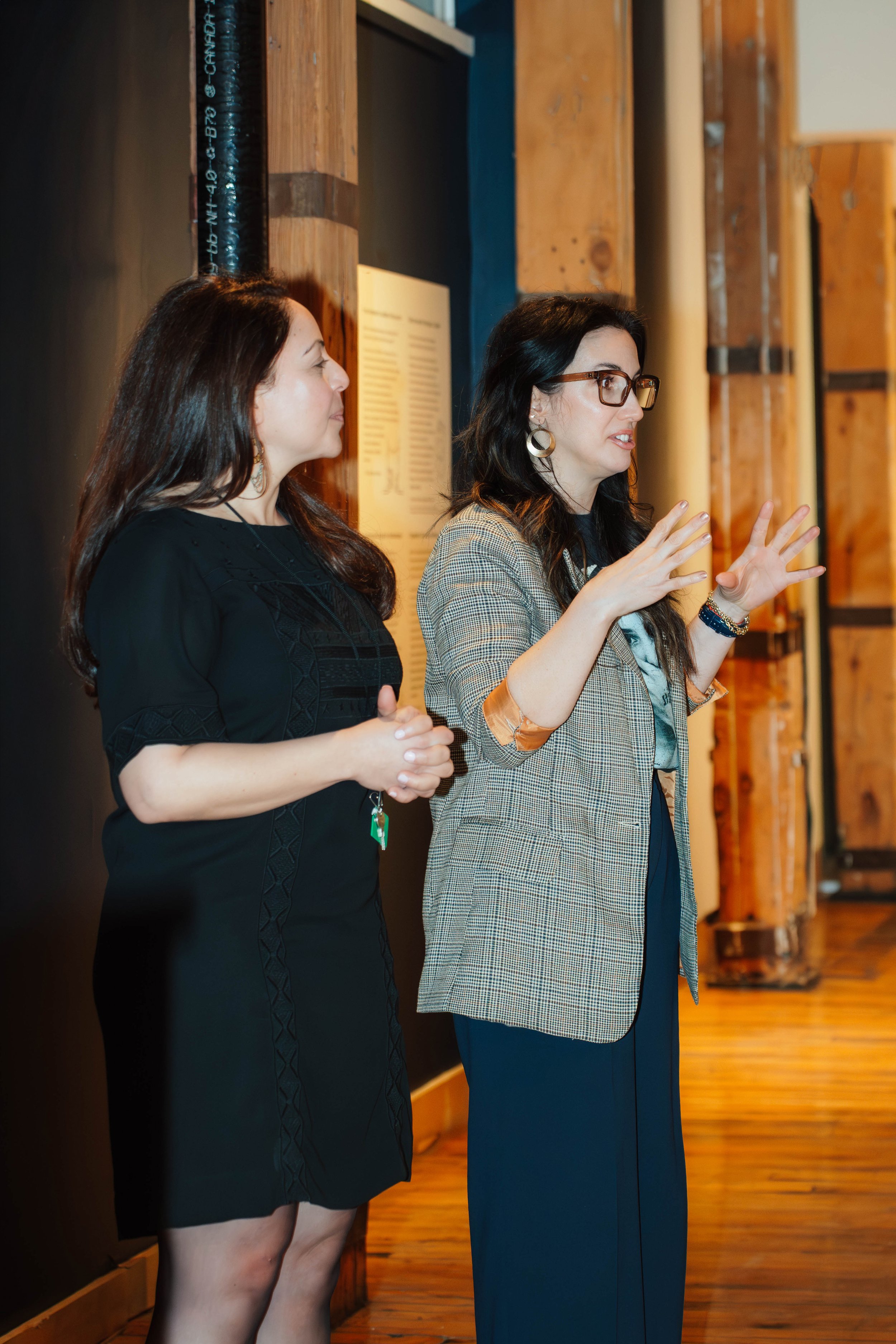 Two women are engaged in a conversation or presentation indoors, standing near a wooden wall with a large pipe overhead. The woman on the right, wearing glasses and earrings, appears to be explaining something with gestures, while the woman on the le