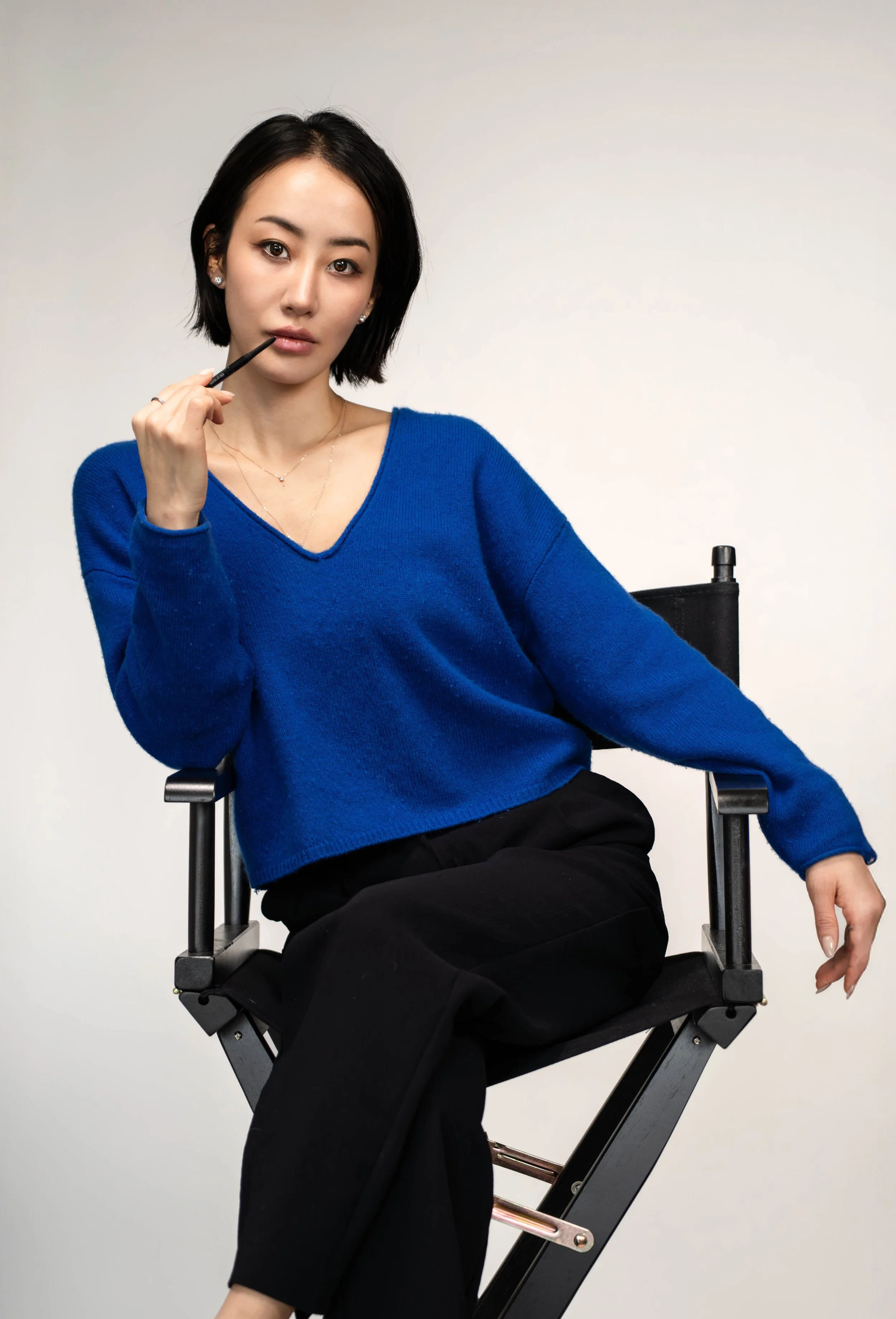 A young woman with short black hair and earrings, wearing a blue V-neck sweater and black pants, sitting on a director's chair, holding a makeup brush near her lips, looking at the camera