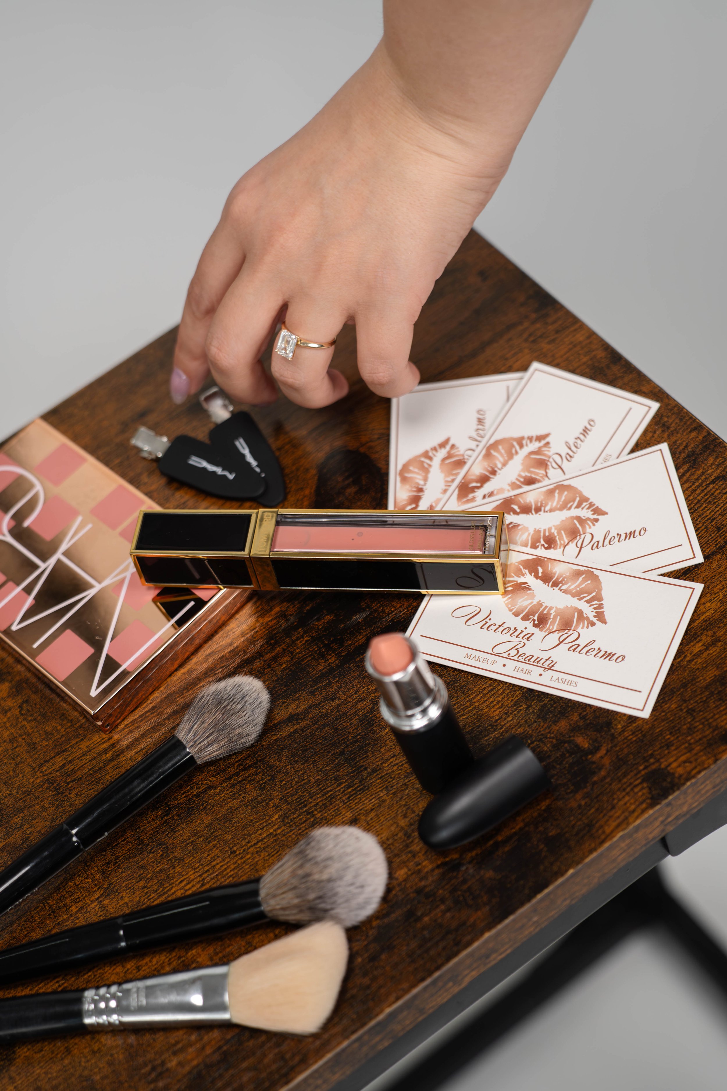 A hand reaching for makeup business cards on a wooden table, with various makeup brushes, lipstick, nail polish, and guitar picks nearby.