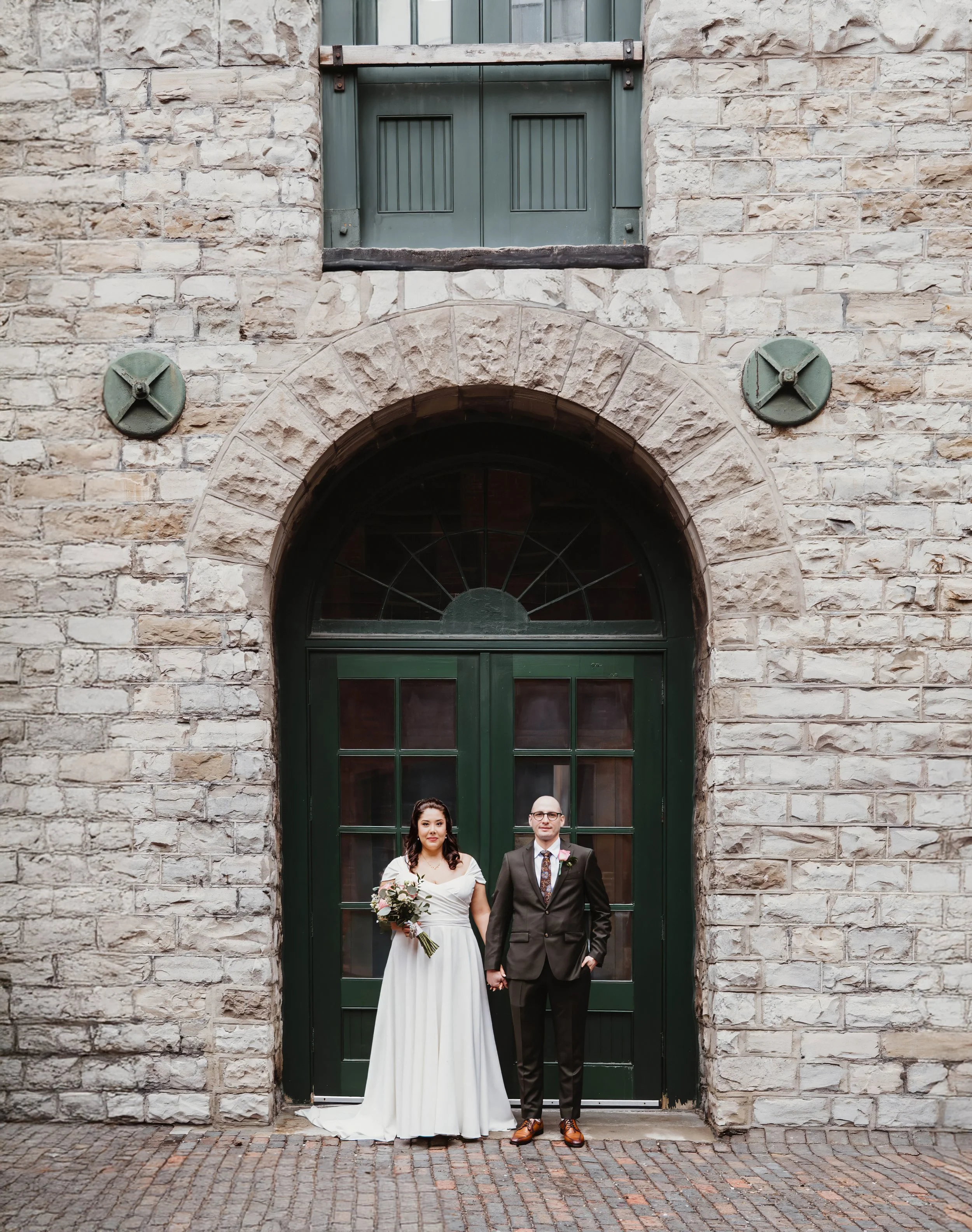 A newlywed couple standing hand in hand in front of a large green door with an arched window, against a stone brick wall.