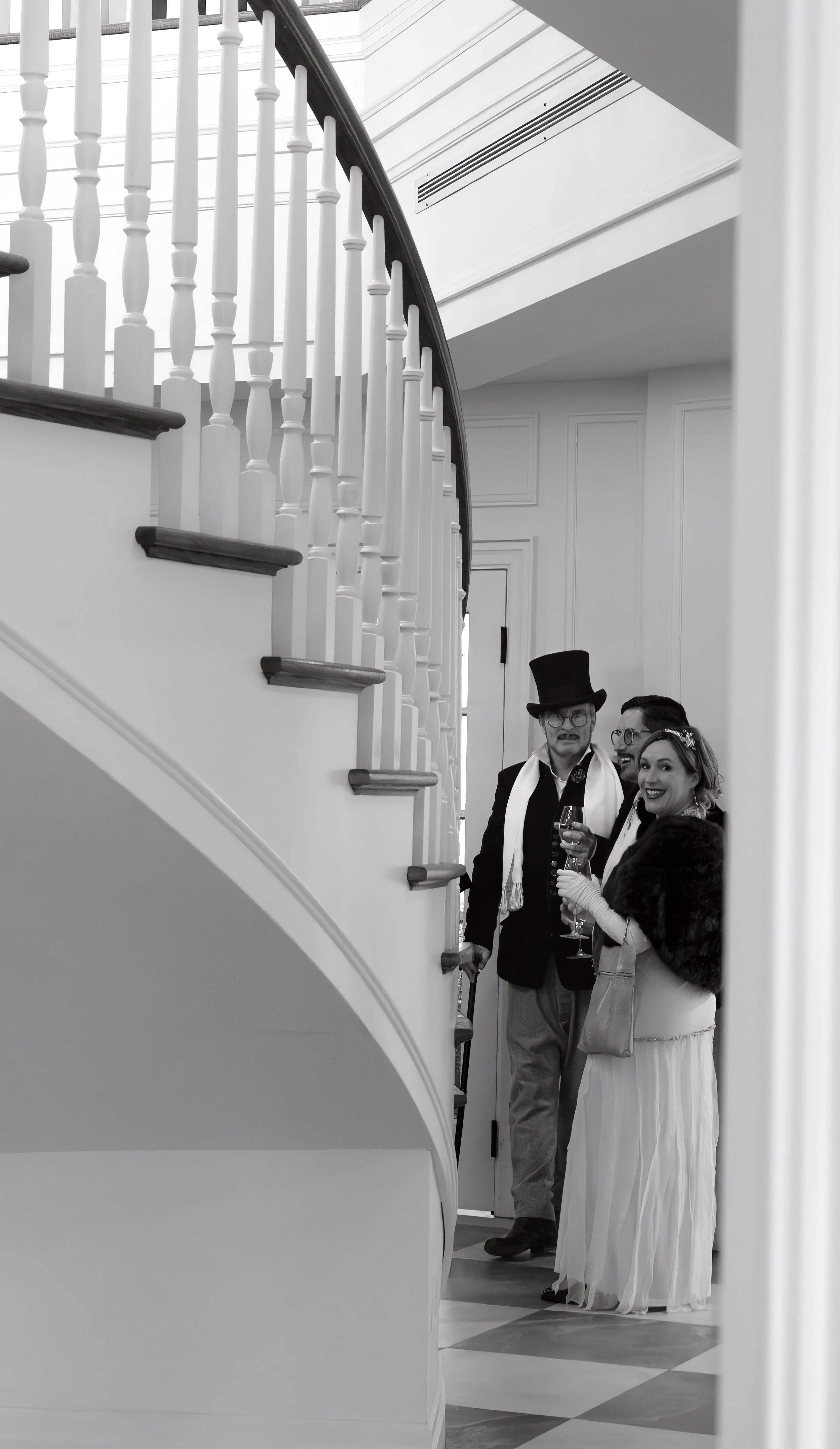 Three people dressed in vintage attire standing next to a staircase, holding glasses of wine, at a formal indoor event.