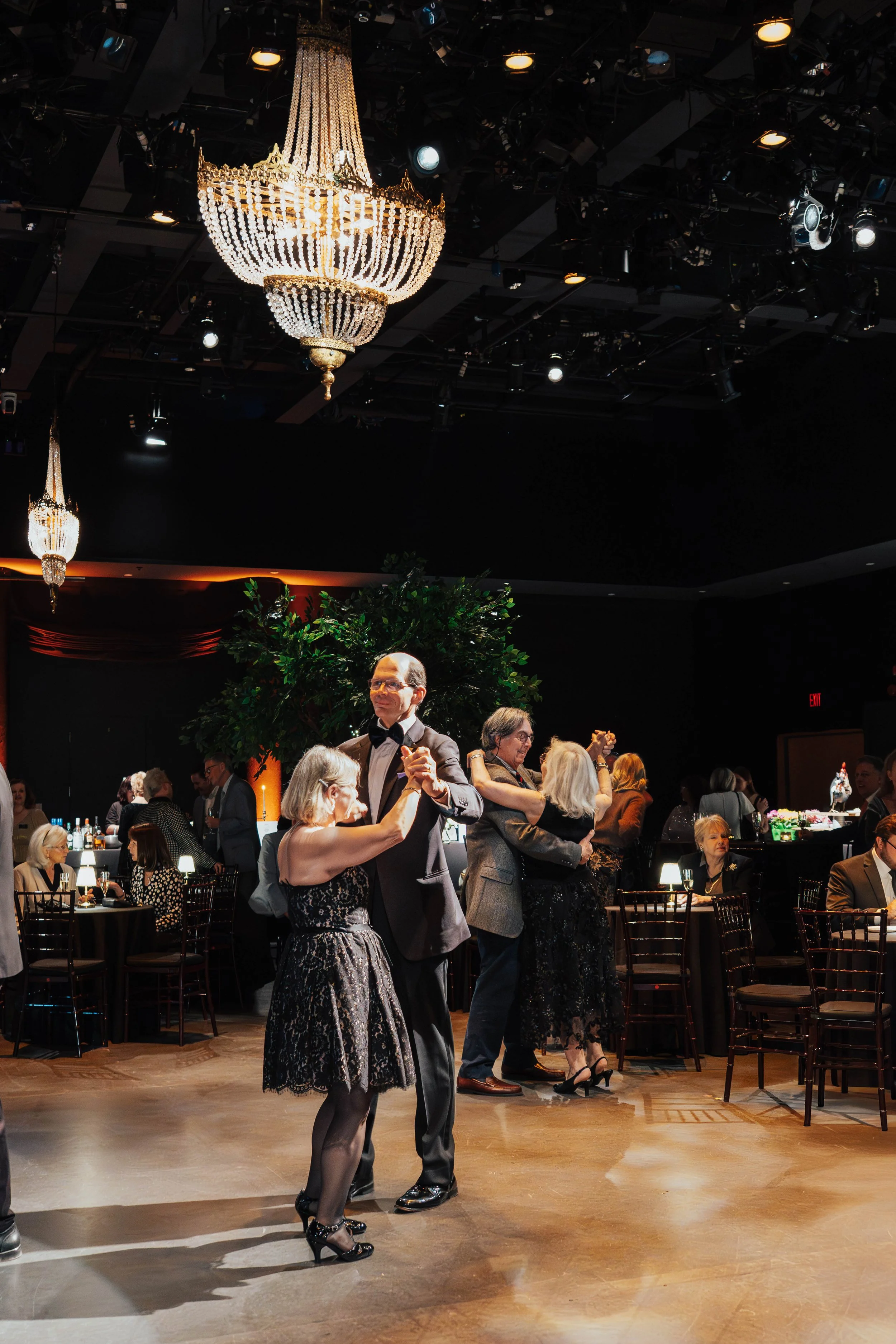People dancing at a formal event in a ballroom with chandeliers and tables in the background.