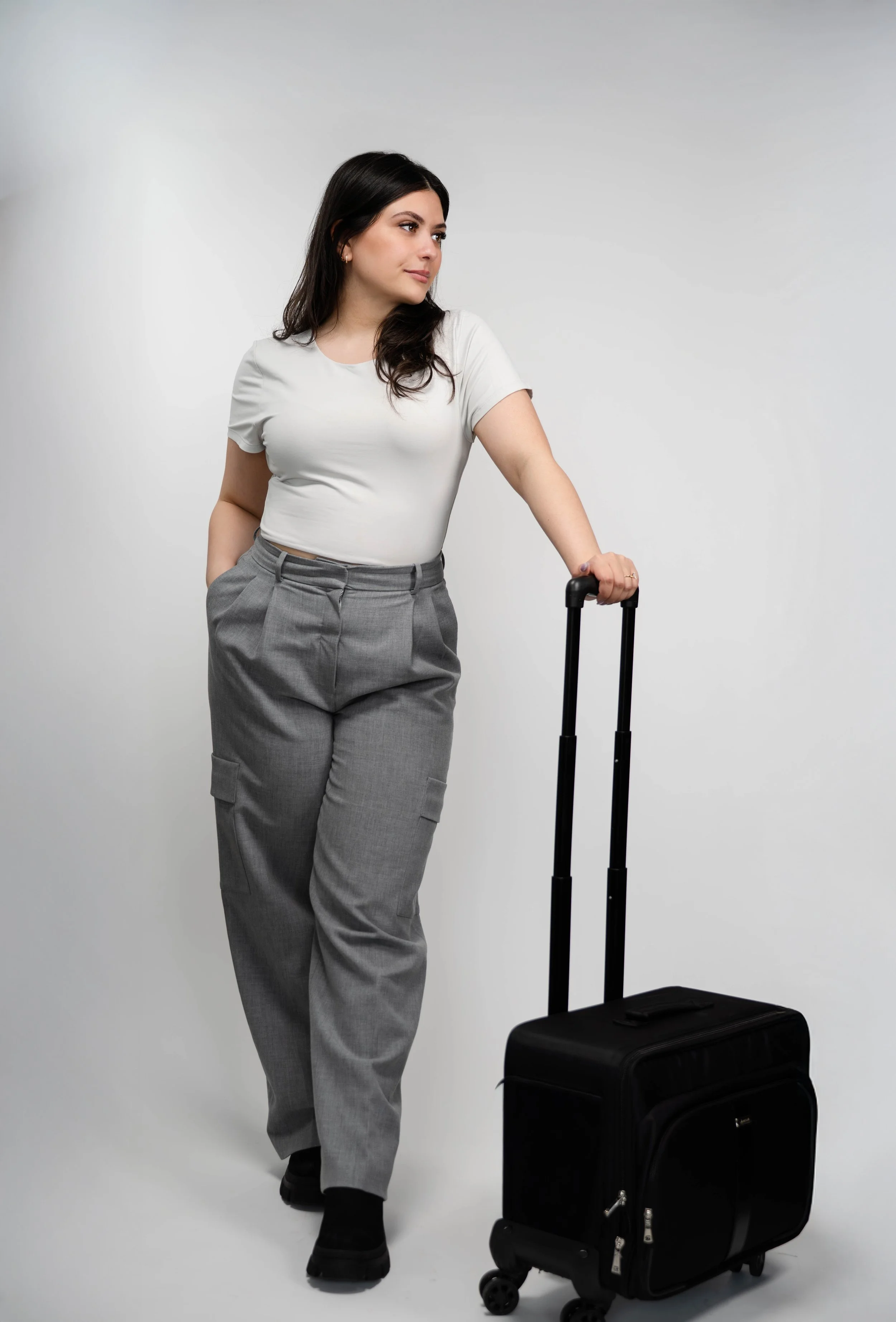 A woman with dark hair wearing a white shirt and gray pants standing with her hand on a rolling black suitcase.
