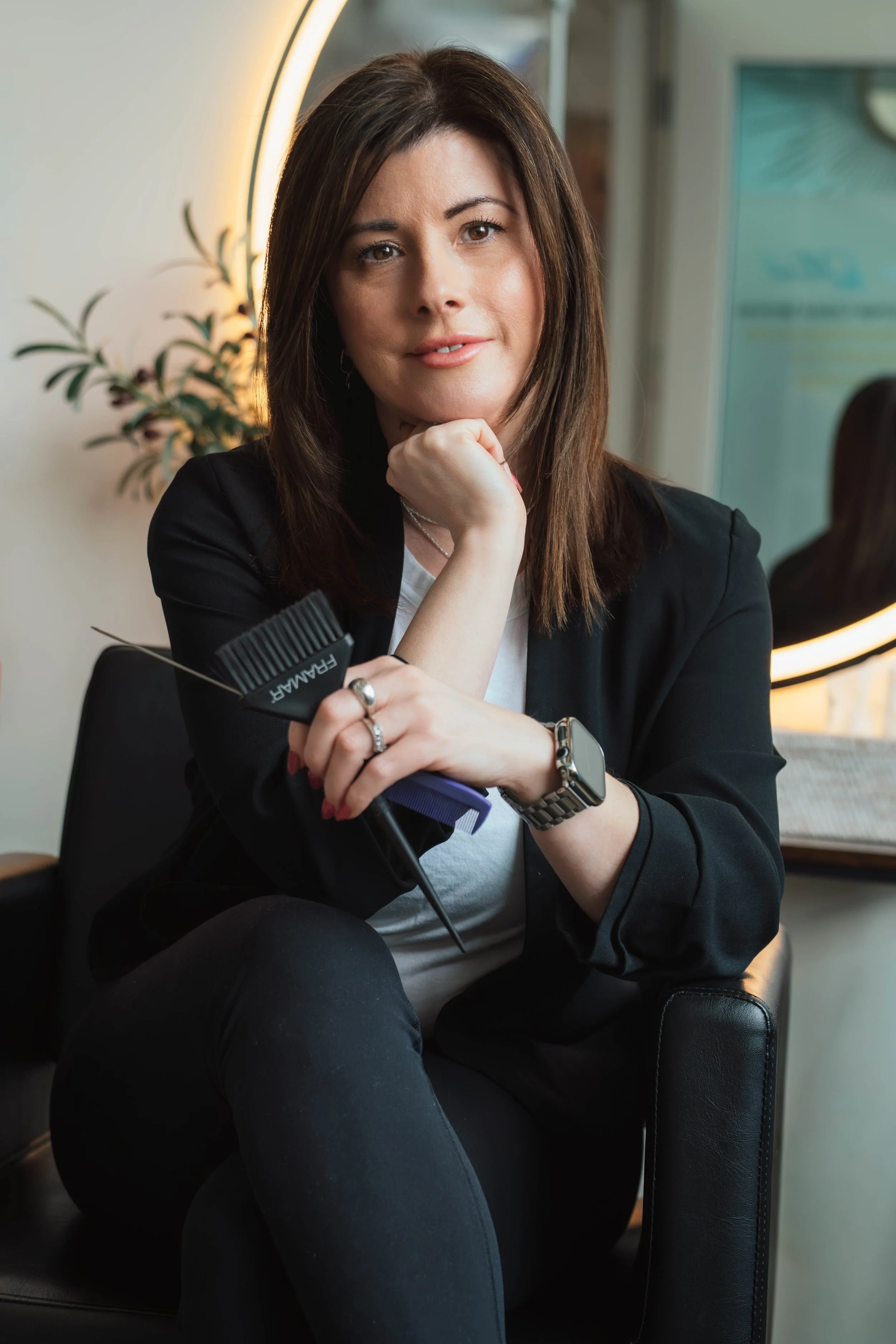 A woman with shoulder-length brown hair, wearing a black blazer, sitting on a black chair in a modern salon or office, holding a hairbrush. She is resting her chin on her hand, looking directly at the camera with a neutral expression, wearing a watch