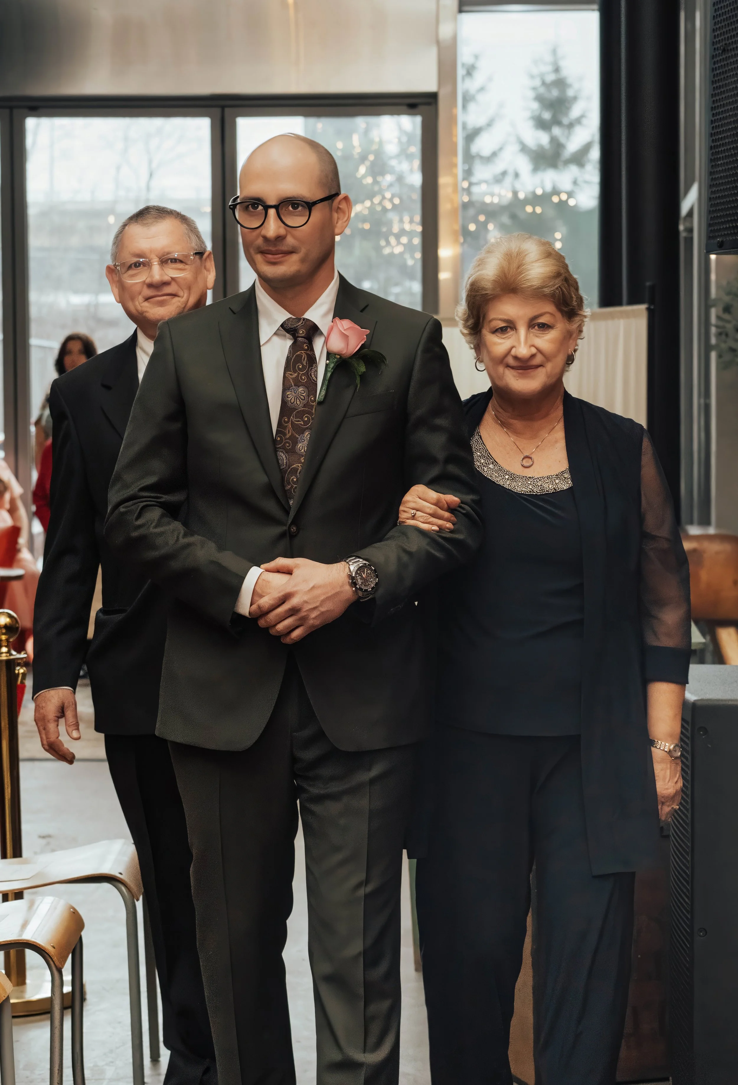 A man in a black suit with glasses and a pink boutonniere holding hands with an older woman in black attire, and an older man in a black suit standing behind them, in a room with large windows and fairy lights.