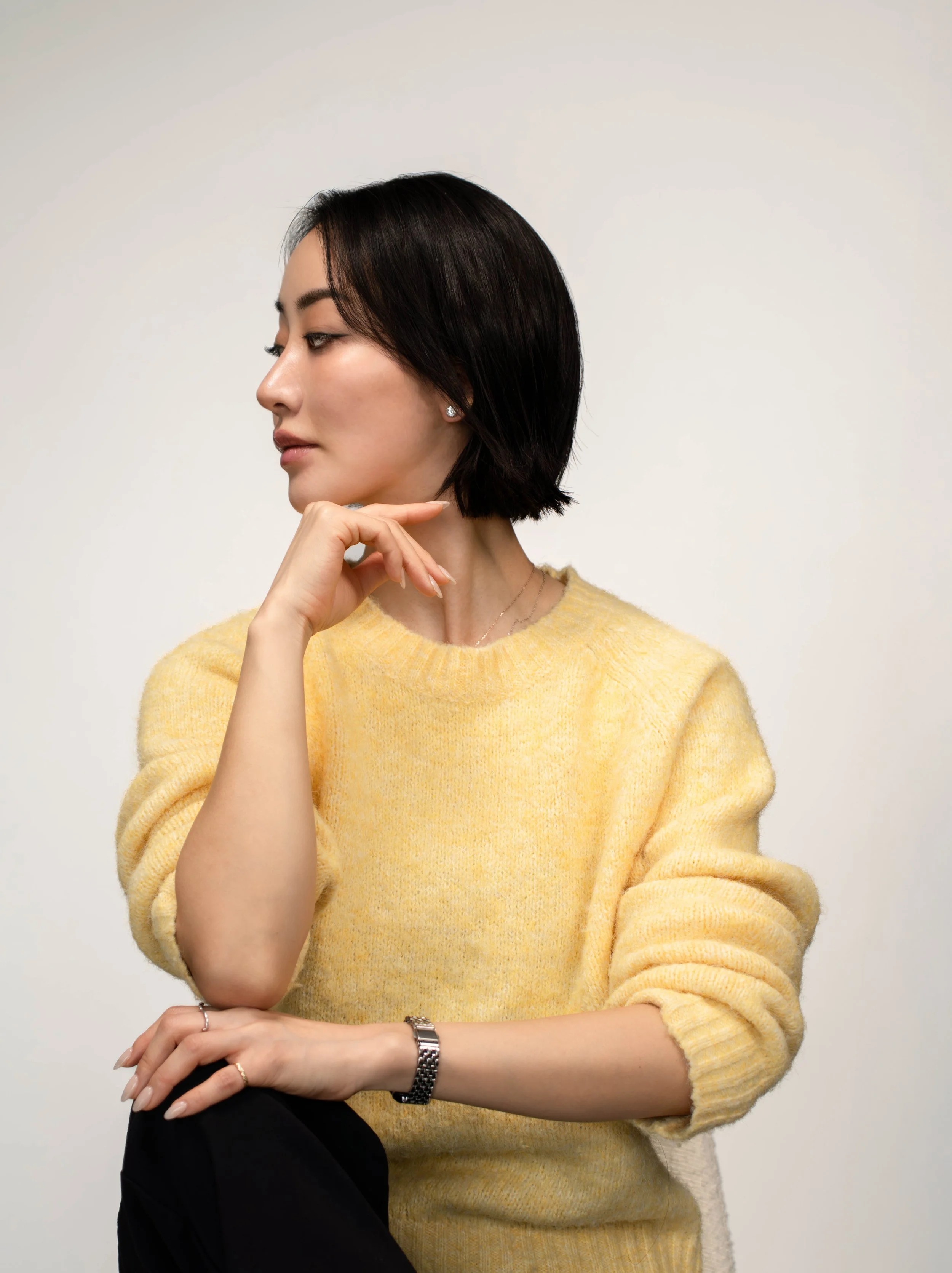 Side profile of a woman with short black hair, wearing a yellow sweater, sitting with her hand resting on her chin against a plain light background.