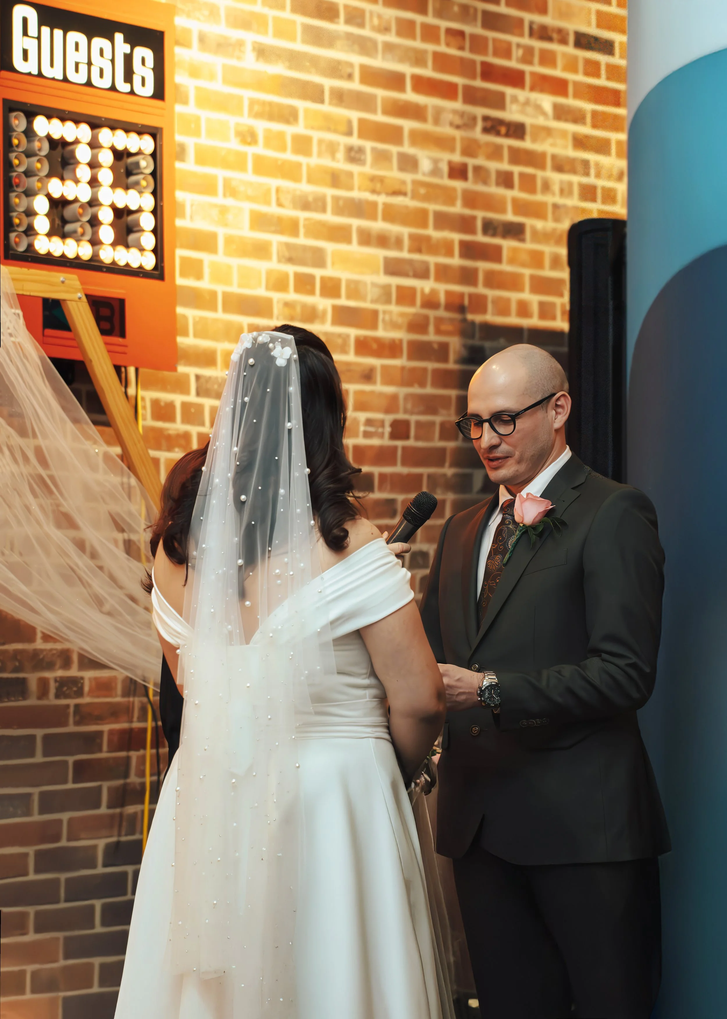 Bride and groom exchanging vows during wedding ceremony, bride in white dress and veil, groom in dark suit with pink boutonniere, brick wall background, person holding microphone, wedding reception setting.