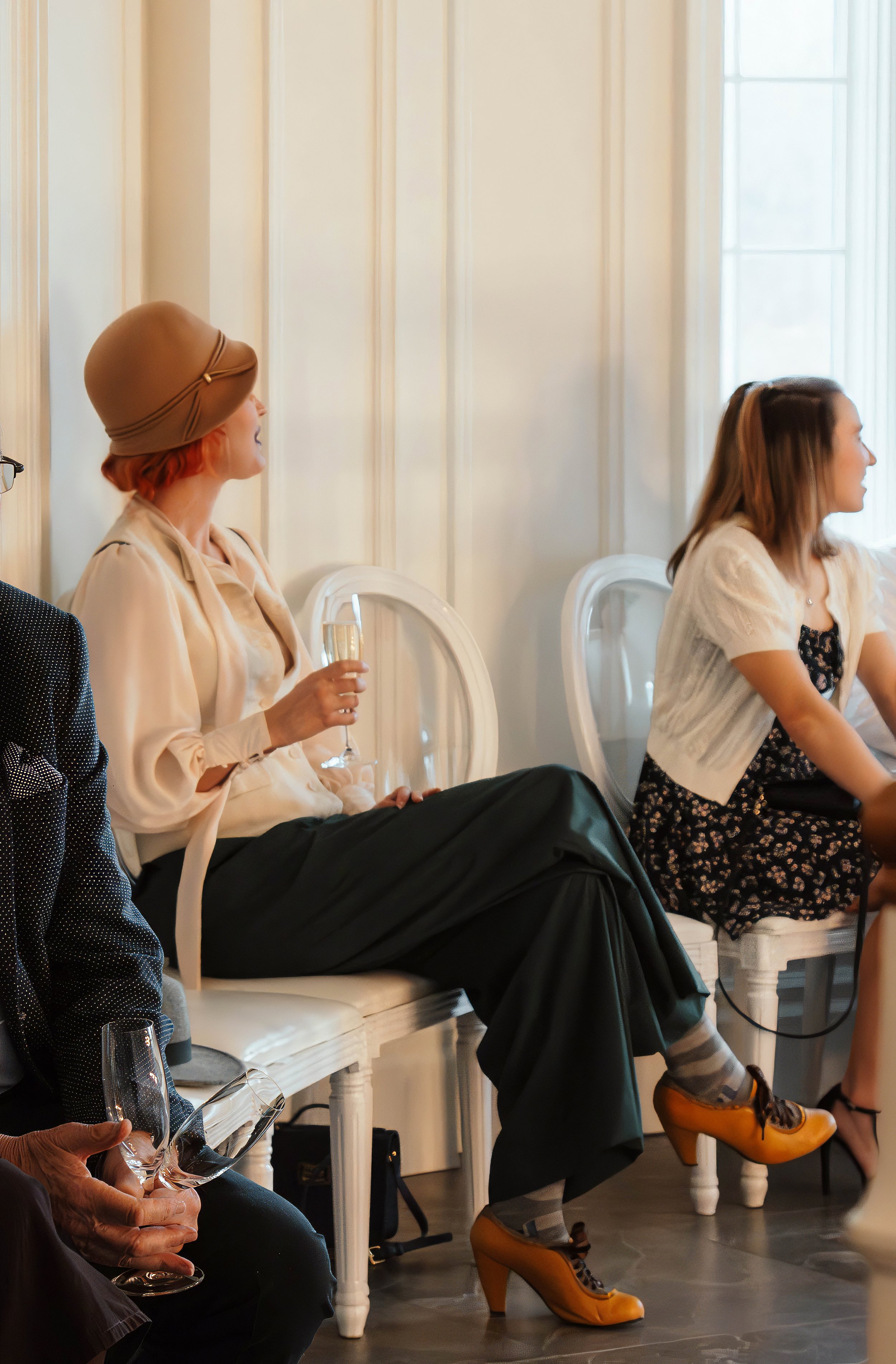 Woman with red hair wearing a beige hat, cream blouse, and black pants sitting on white chair, holding a glass of champagne at a social event.