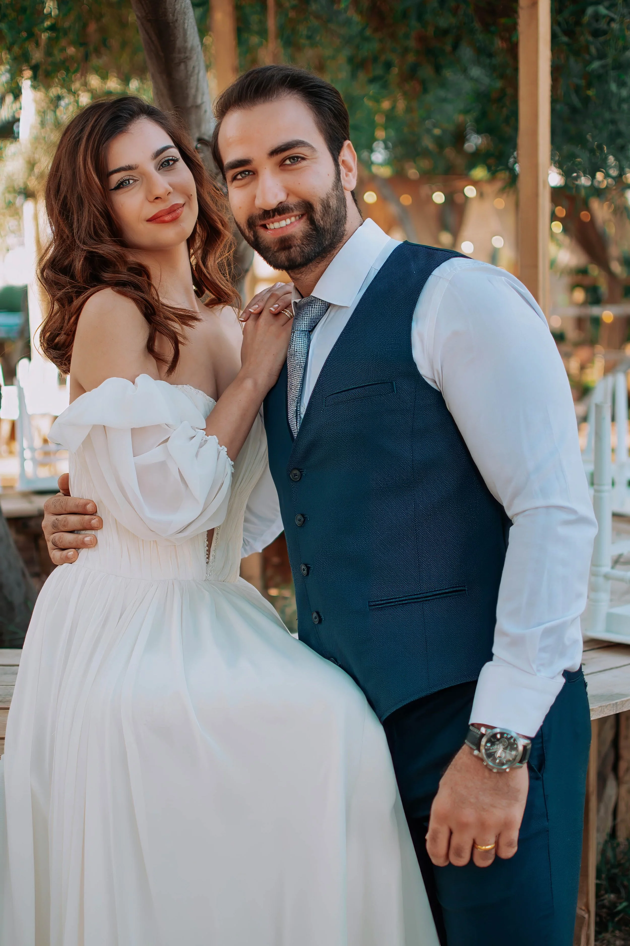 A smiling couple dressed in wedding attire, outdoors with trees and blurred lights in the background.