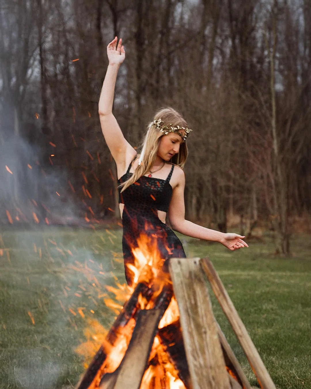 A young woman with long blonde hair wearing a black dress and a flower crown, dancing near a bonfire outdoors on a grassy field with bare trees in the background.