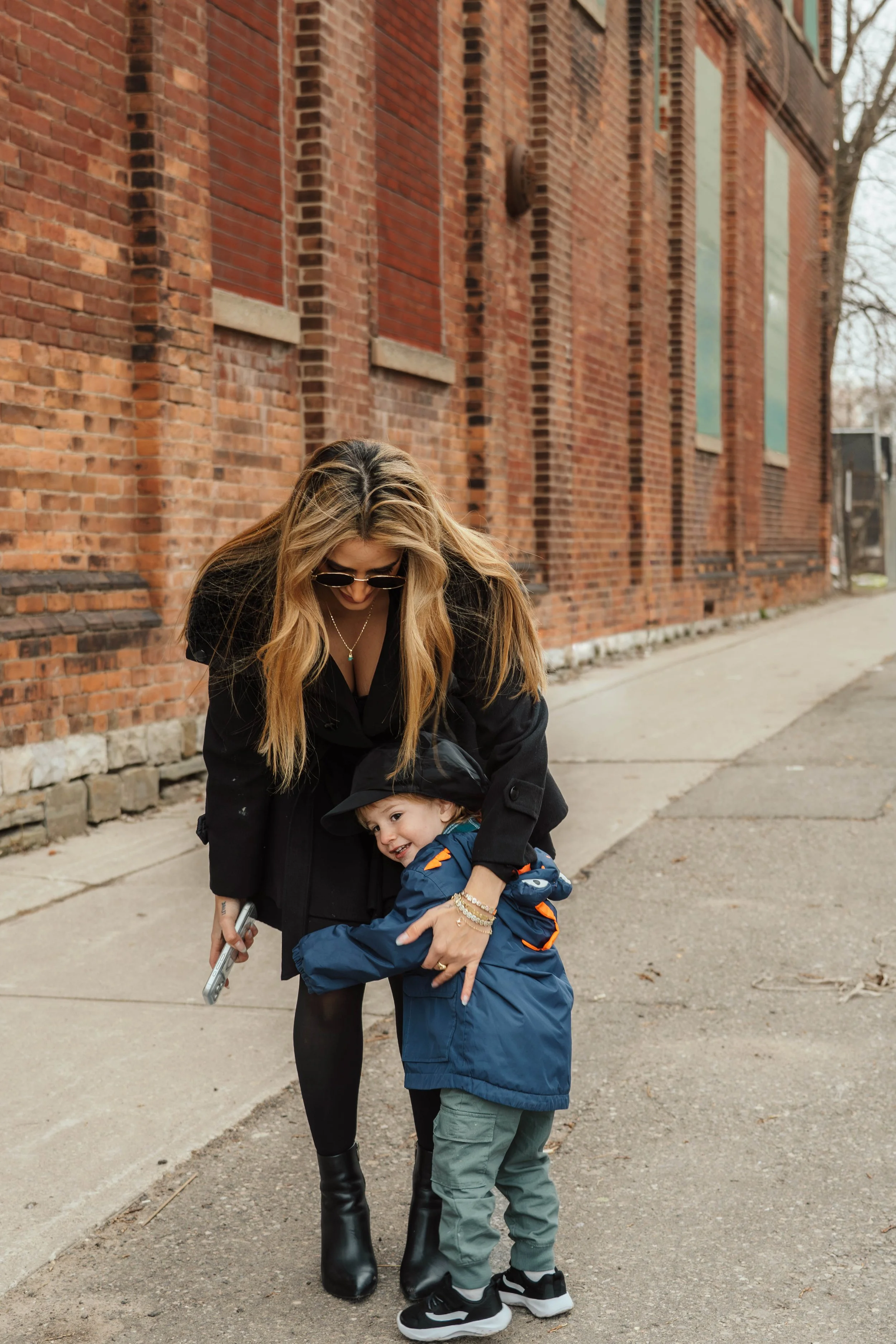 A woman with long blonde hair, wearing sunglasses, black coat, and black boots, is hugging a young boy in a blue jacket, green pants, and black sneakers, on a sidewalk beside a brick building.