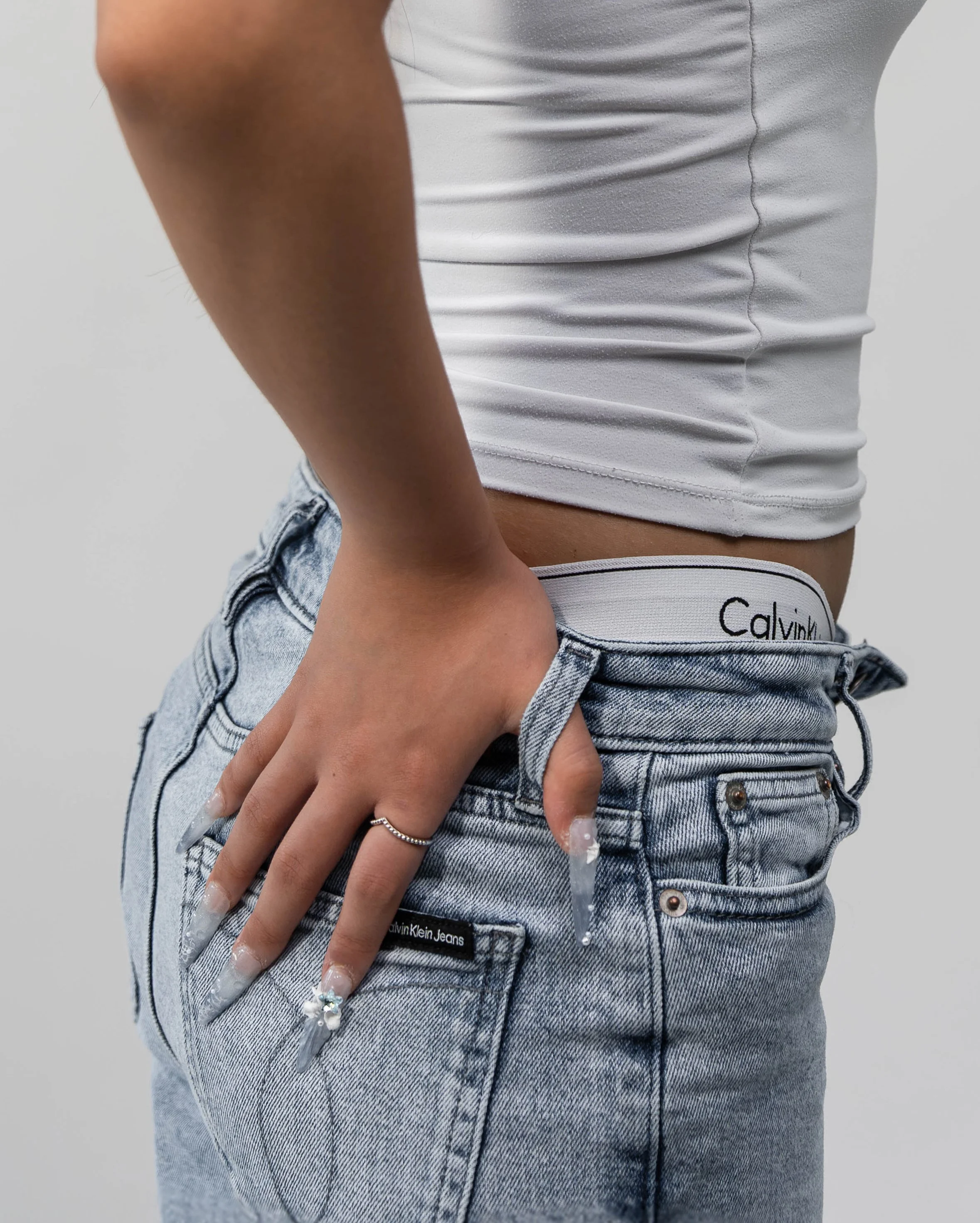 Close-up of a person wearing a white tank top and light-wash Calvin Klein jeans, with their hand resting on their hip, against a plain background.