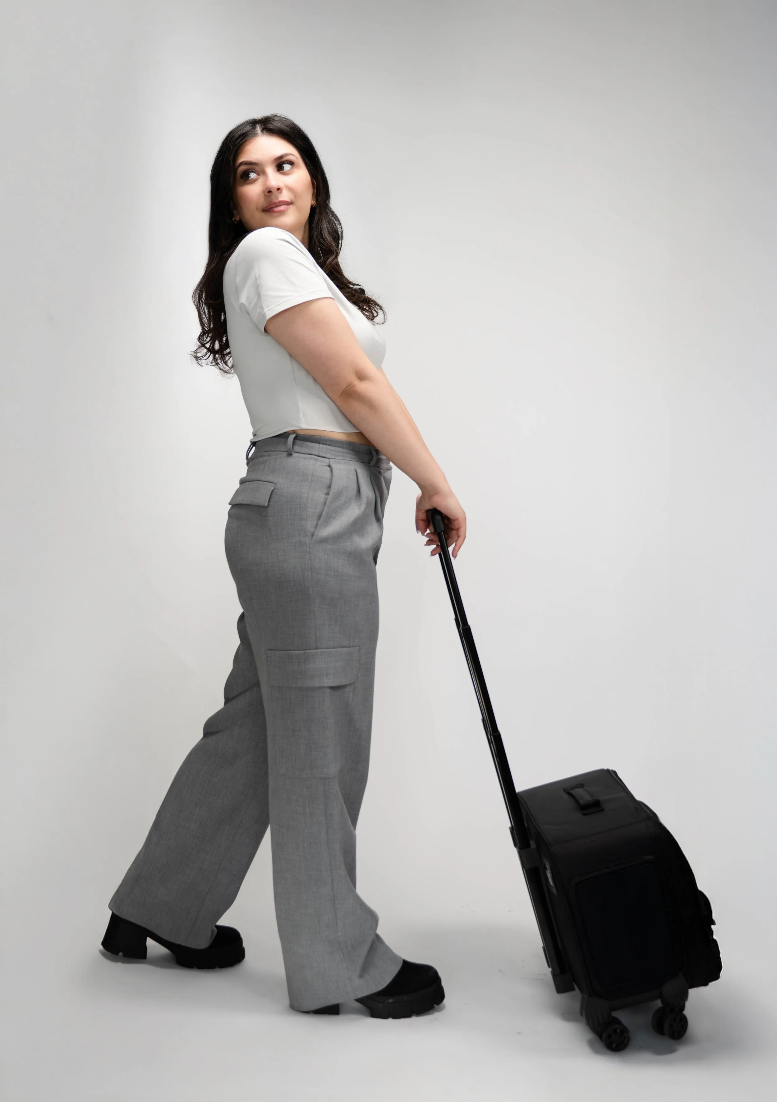 A woman with dark hair wearing a white t-shirt and gray wide-leg pants stands with her hand on a rolling black suitcase, looking to the side against a plain white background.