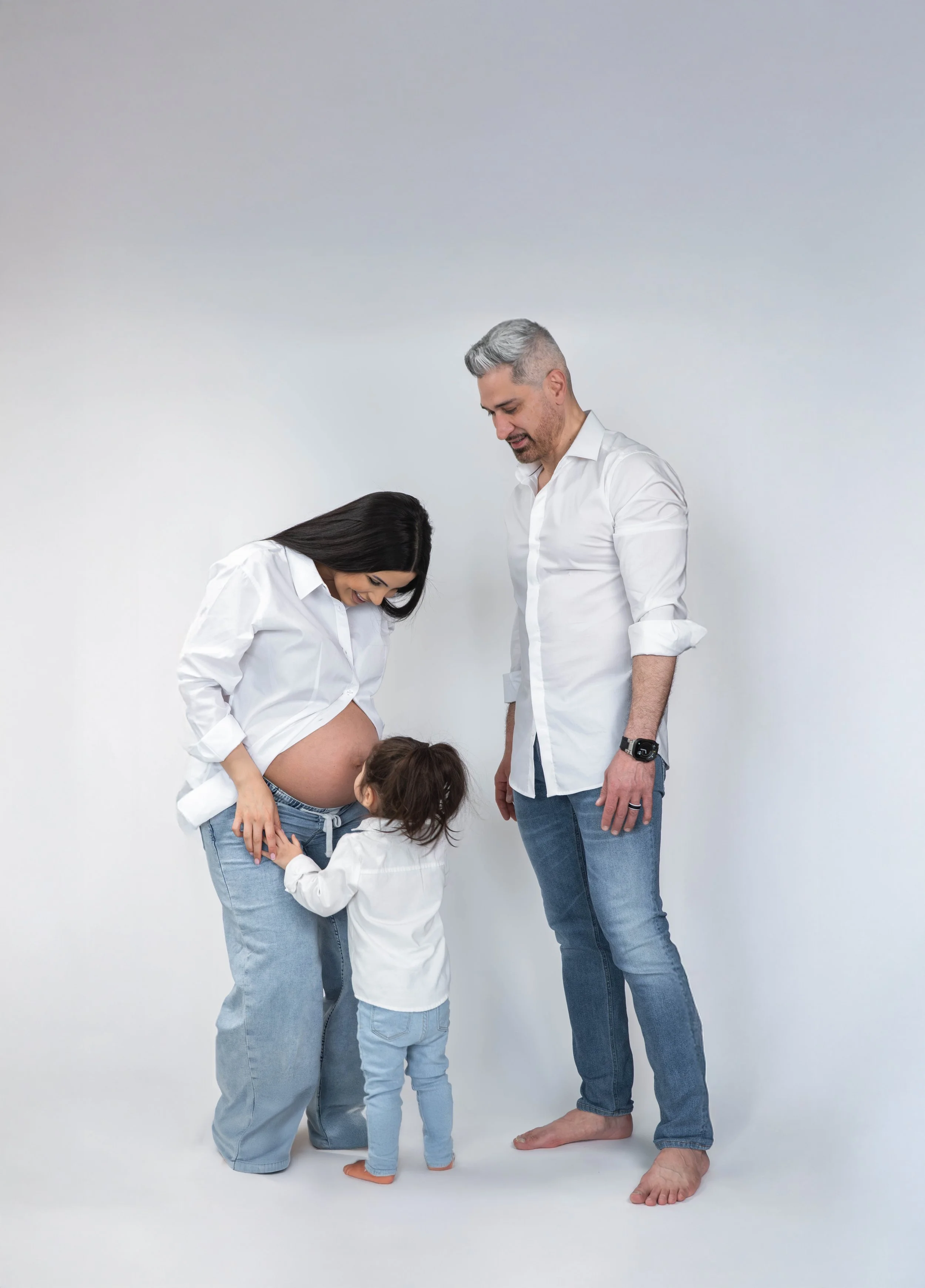 A pregnant woman with long dark hair, a little girl, and a man with gray hair, all in casual white shirts and jeans, standing against a plain white background. The woman is smiling, touching her belly, and looking down at it, while the man and girl a