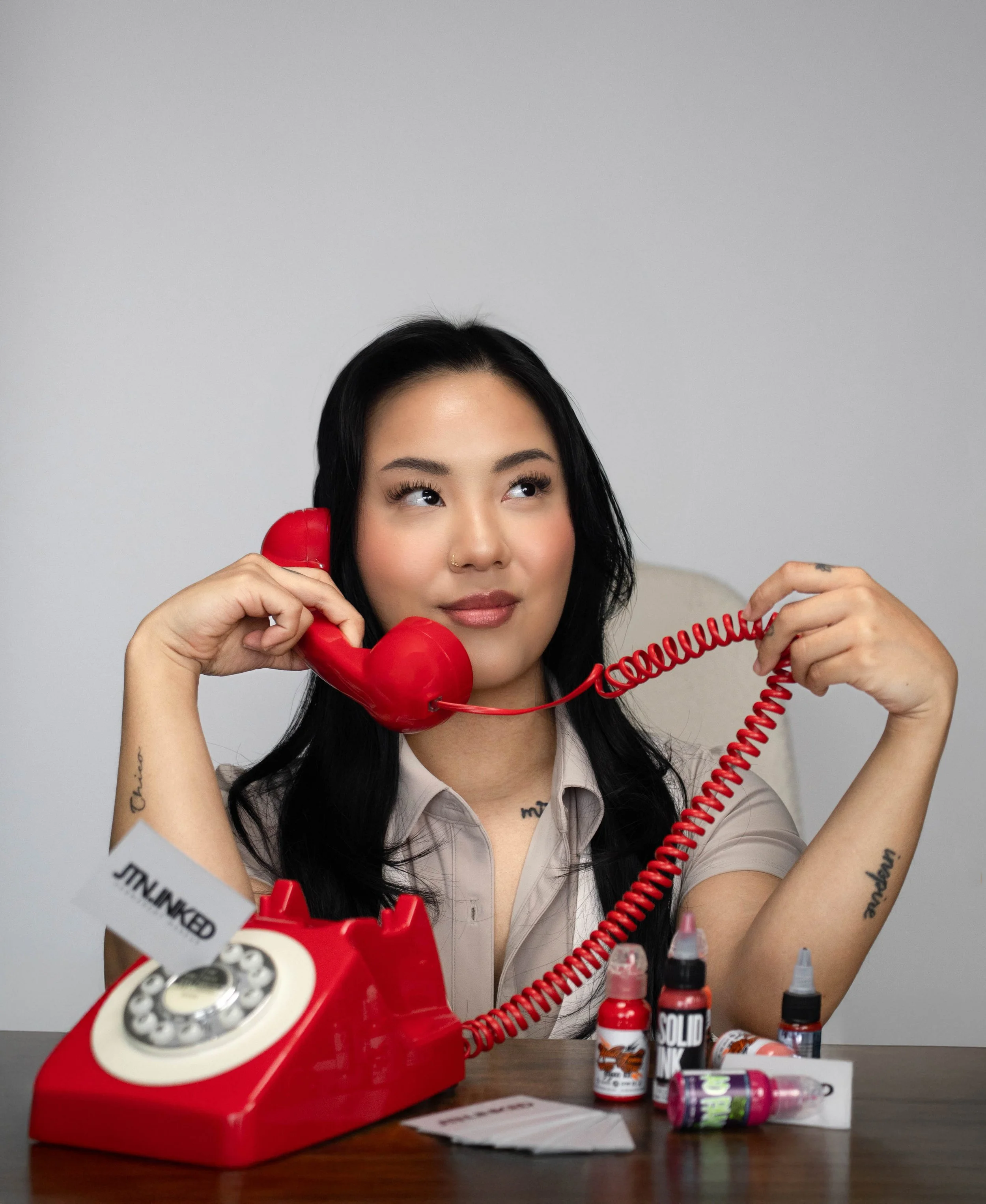 A woman with long black hair and tattoos on her arms holding a red rotary phone to her ear, with various ink bottles and a piece of paper on the table in front of her.