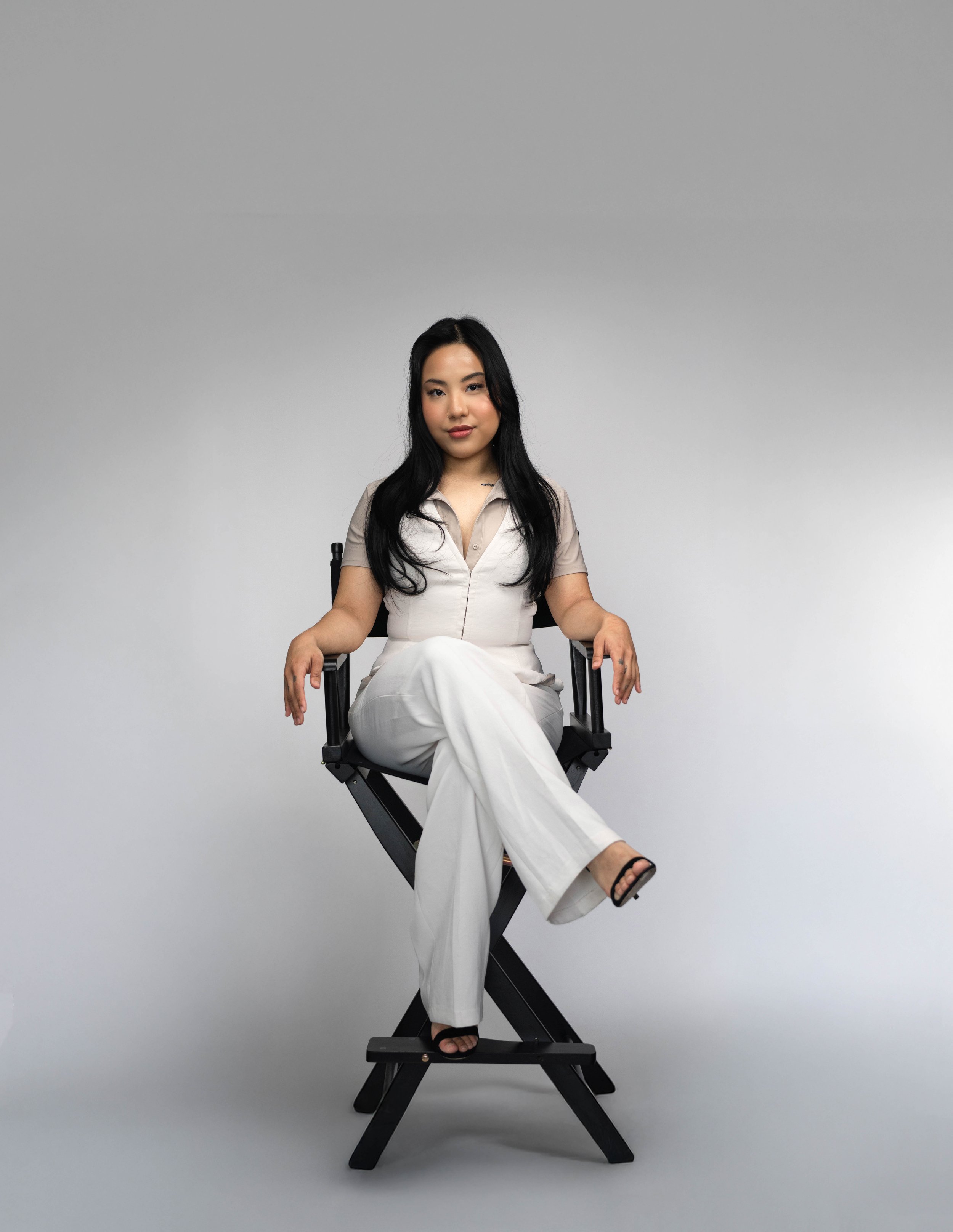 A woman with long black hair sitting cross-legged on a director's chair against a plain gray background, wearing a beige short-sleeve top and white pants.