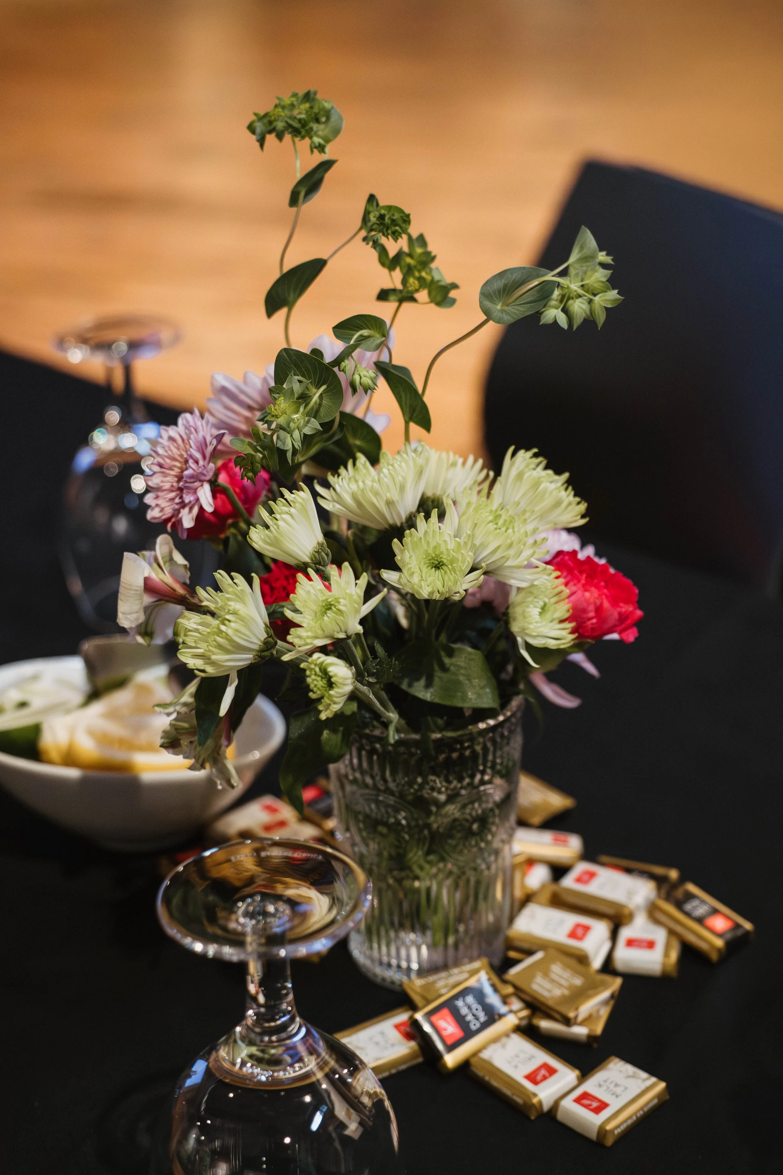 A flower arrangement with pink, white, and red flowers in a glass vase on a black table, surrounded by scattered chocolate bars, wine glasses, and a white bowl with food, with a blurry background.