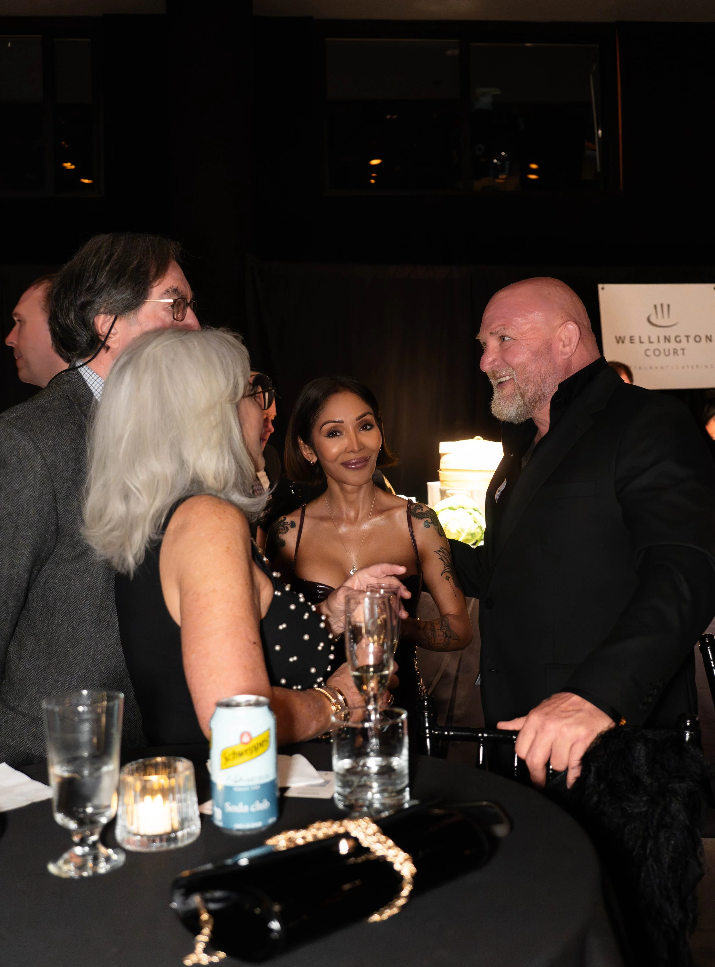 Group of people socializing at a formal event, including a woman with gray hair and glasses, a woman with tattoos wearing a black dress, and a bearded man in a black suit. There are drinks and a small candle on the table in front of them.