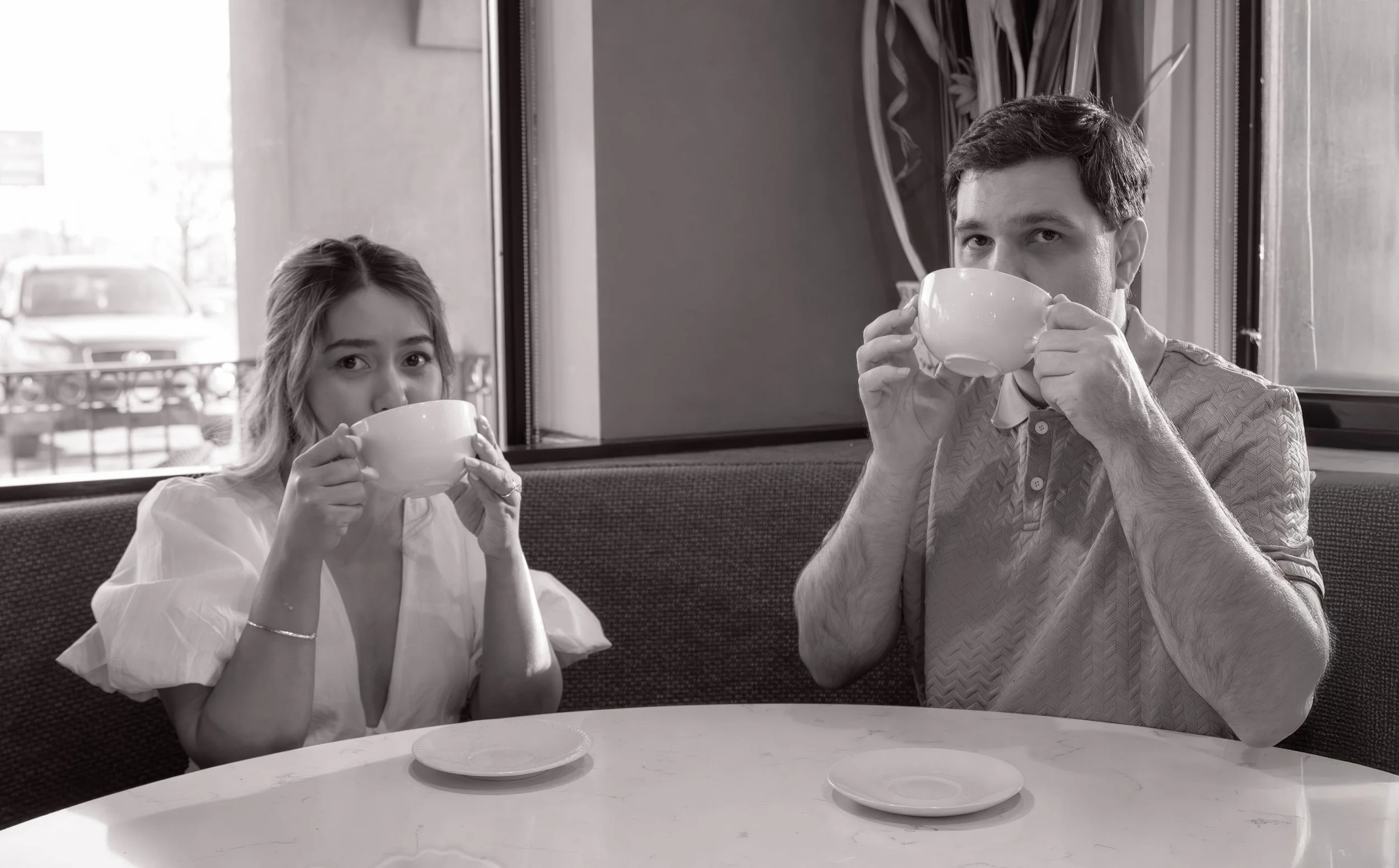 A man and woman sitting at a diner table drinking from white cups, with a window showing a car outside.