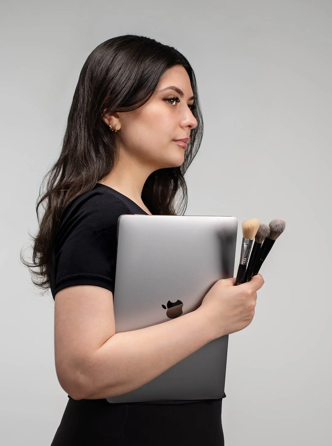 Woman with dark hair holding a silver MacBook and makeup brushes, wearing a black top, against a plain background.
