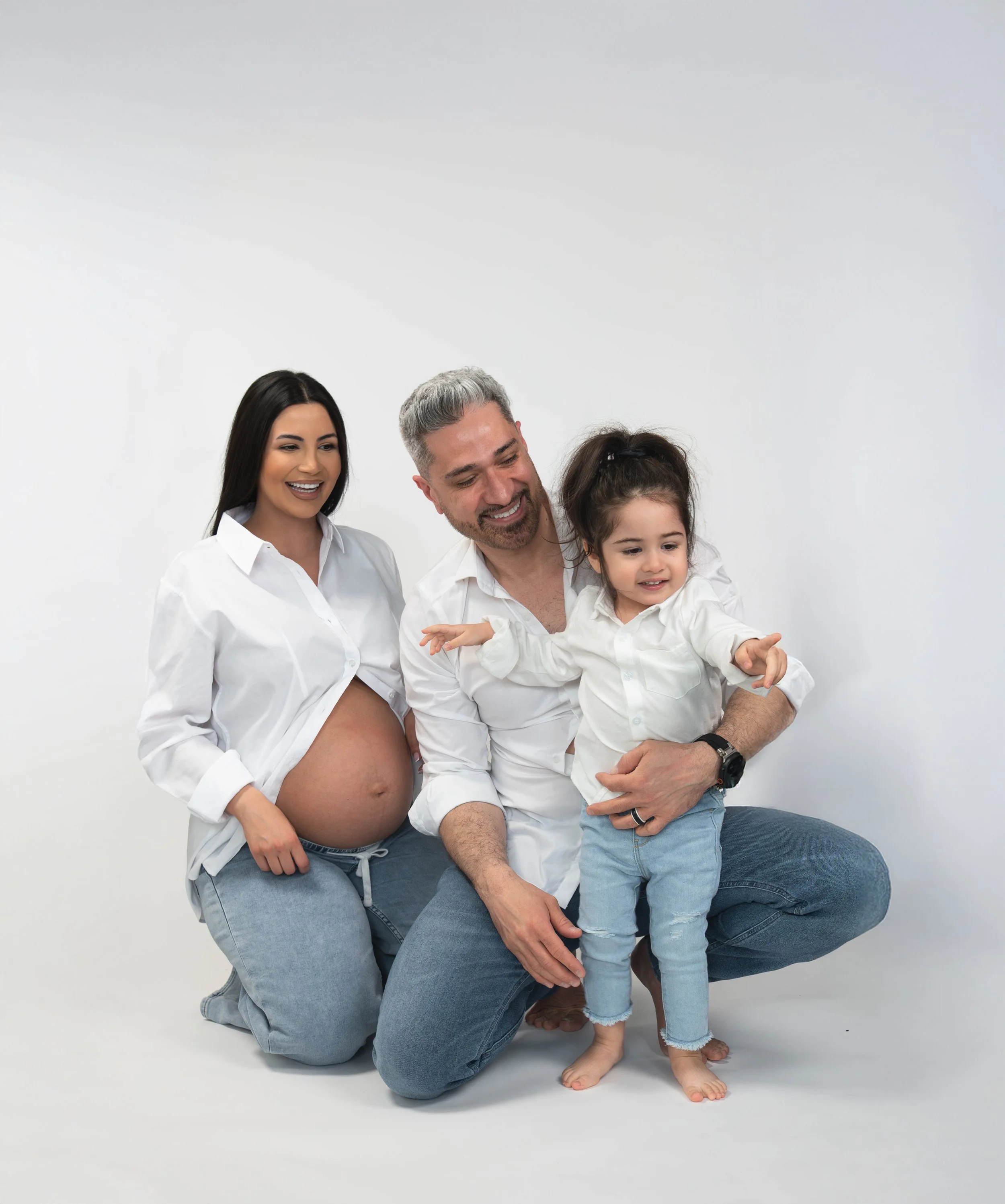A diverse family of four, including a pregnant woman, a man, a young girl, and a woman with curly hair, dressed in white shirts and jeans, smiling and with a plain white background.