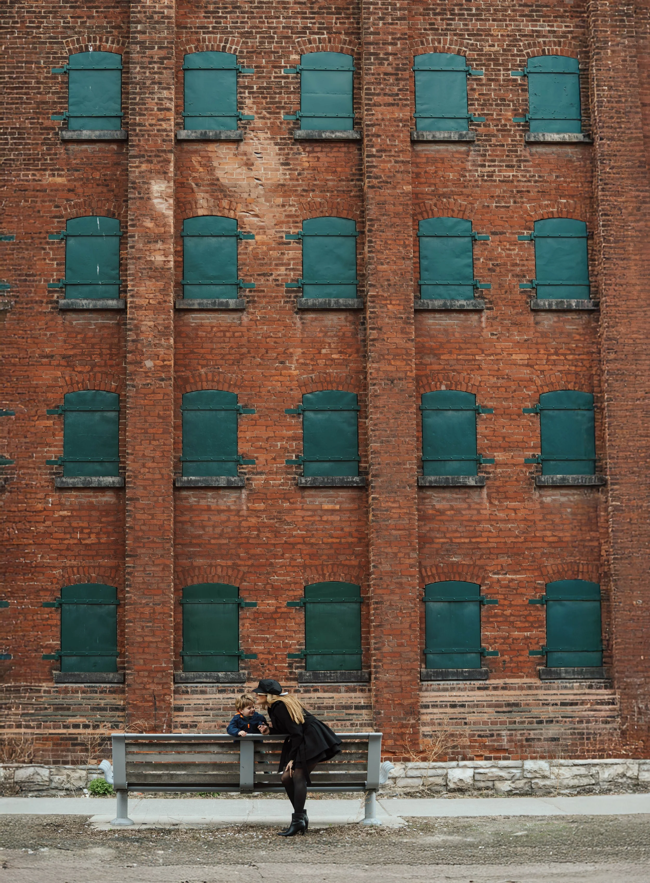 A woman and a child sitting on a bench in front of a brick building with green shutters, with the woman leaning towards the child, during daytime.