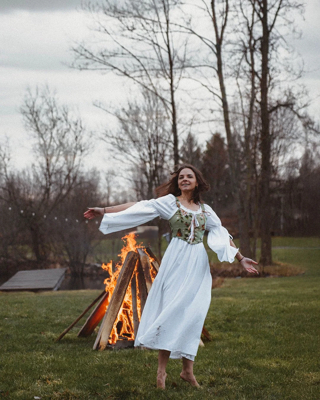 A woman in a white dress and a folk-inspired top dancing barefoot outdoors near a bonfire, with bare trees in the background on a cloudy day.