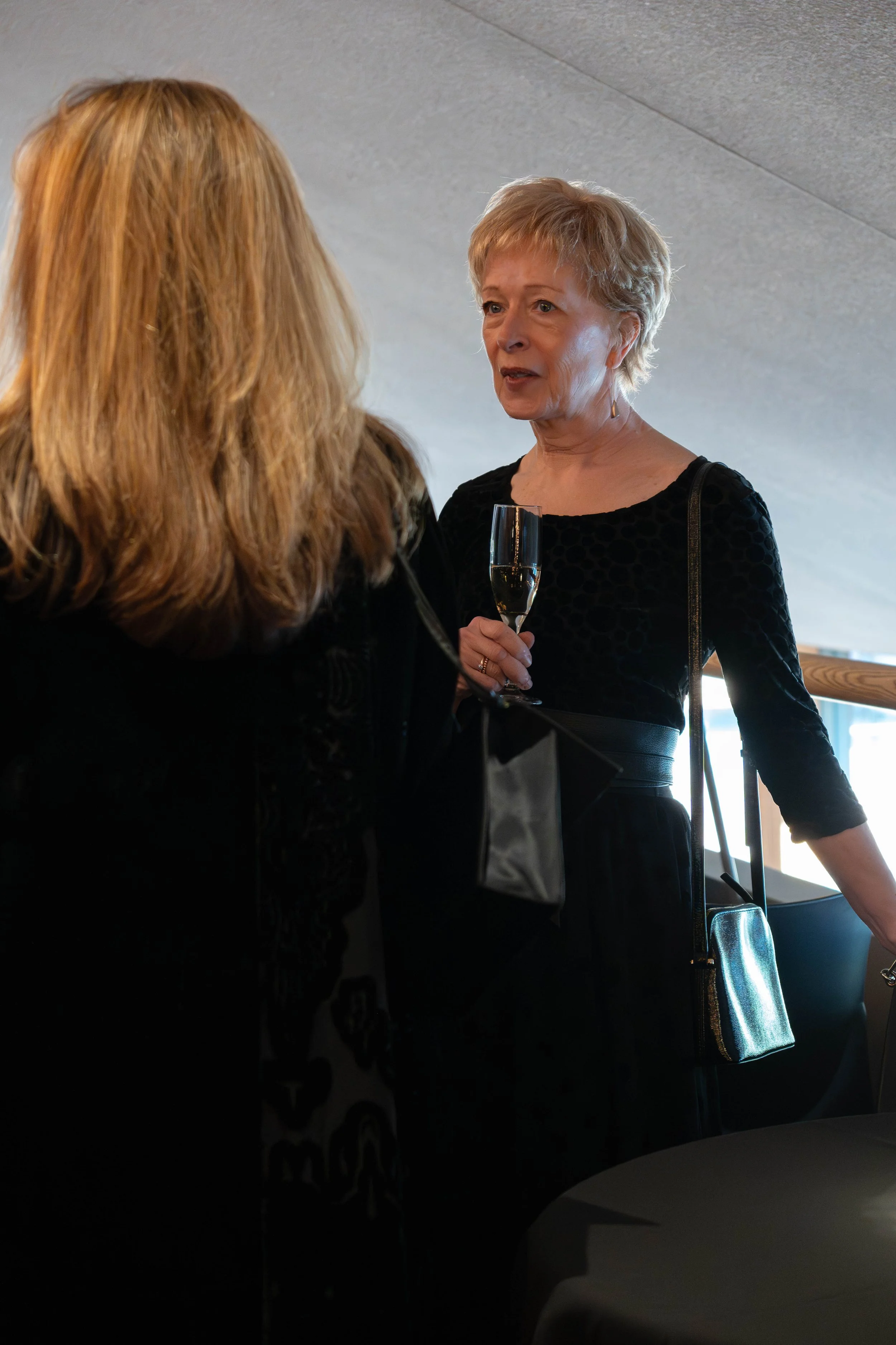 Two women are having a conversation at an indoor social event, one is holding a glass of champagne.