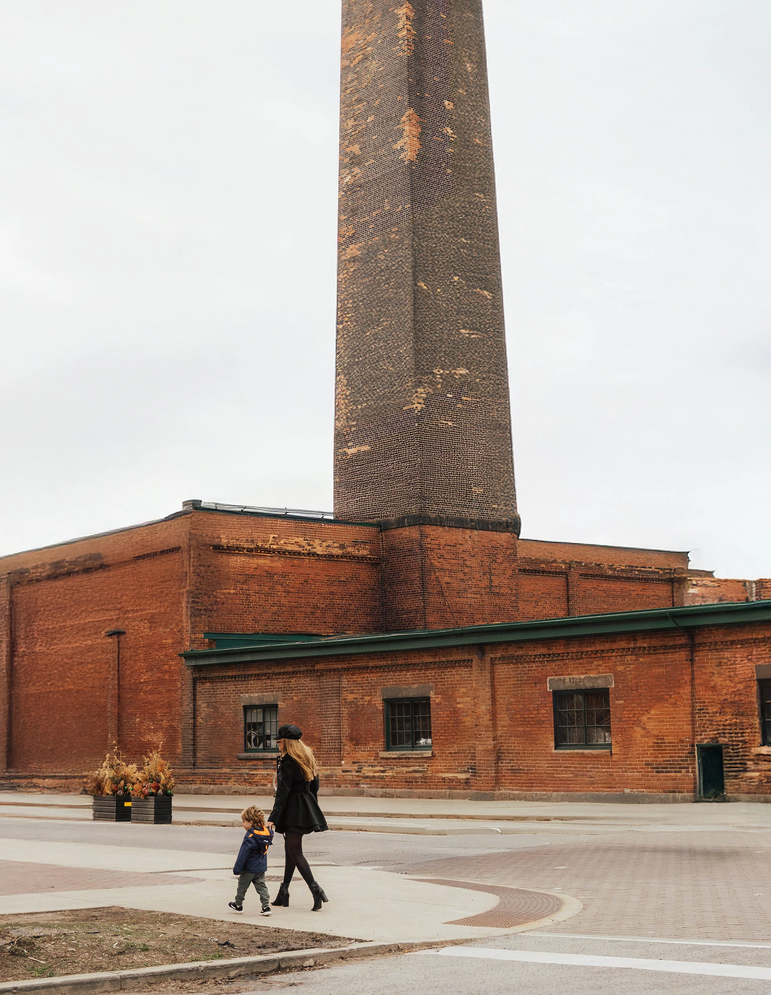 A woman walking with two children across a street in front of a large red brick industrial building with a prominent chimney against an overcast sky.