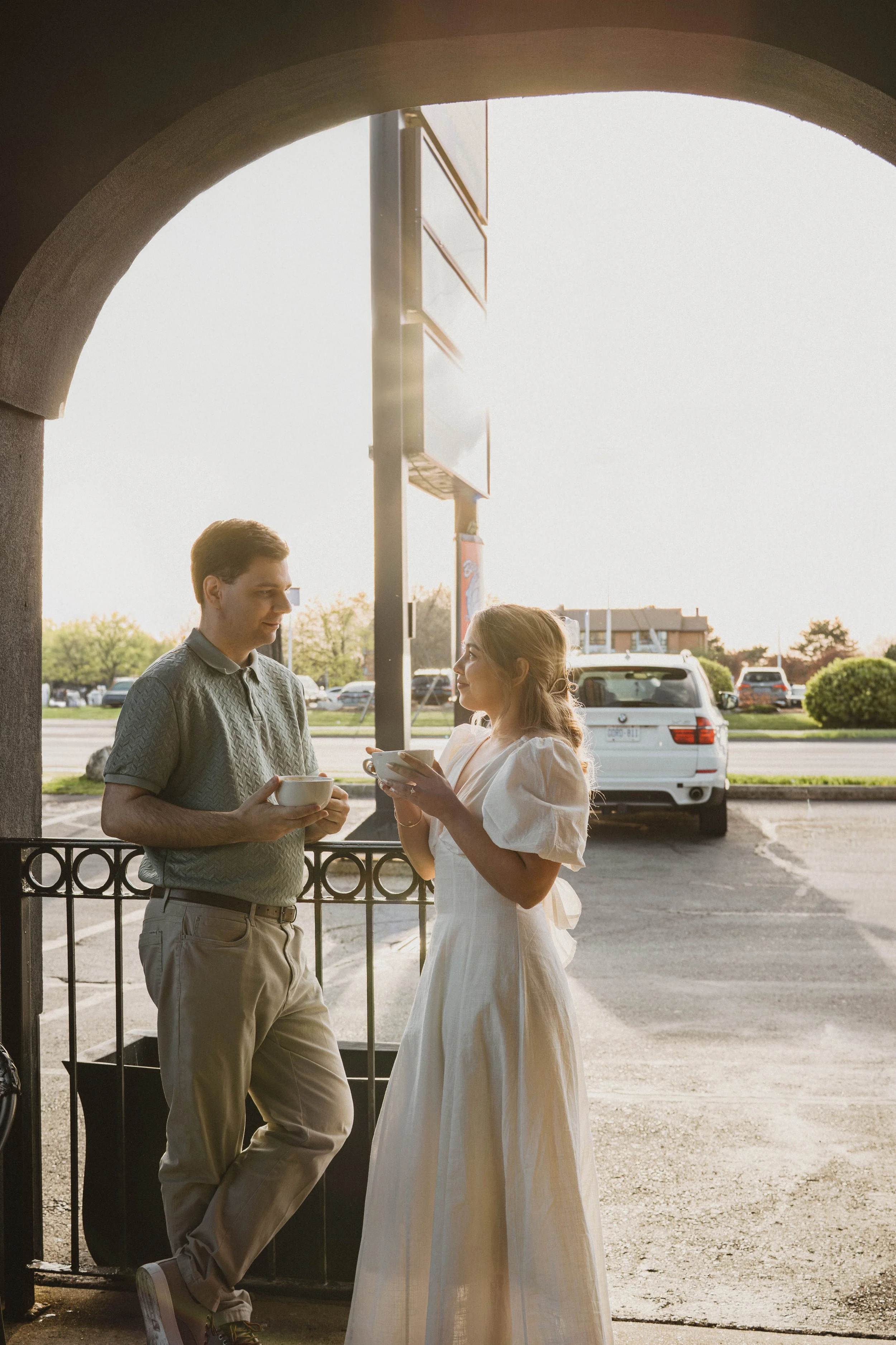 A man and woman standing outside having a conversation. The man is holding a bowl and spoon, and the woman is holding a bowl with a spoon. They are under an archway near parked cars, with a building and greenery in the background, during what appears