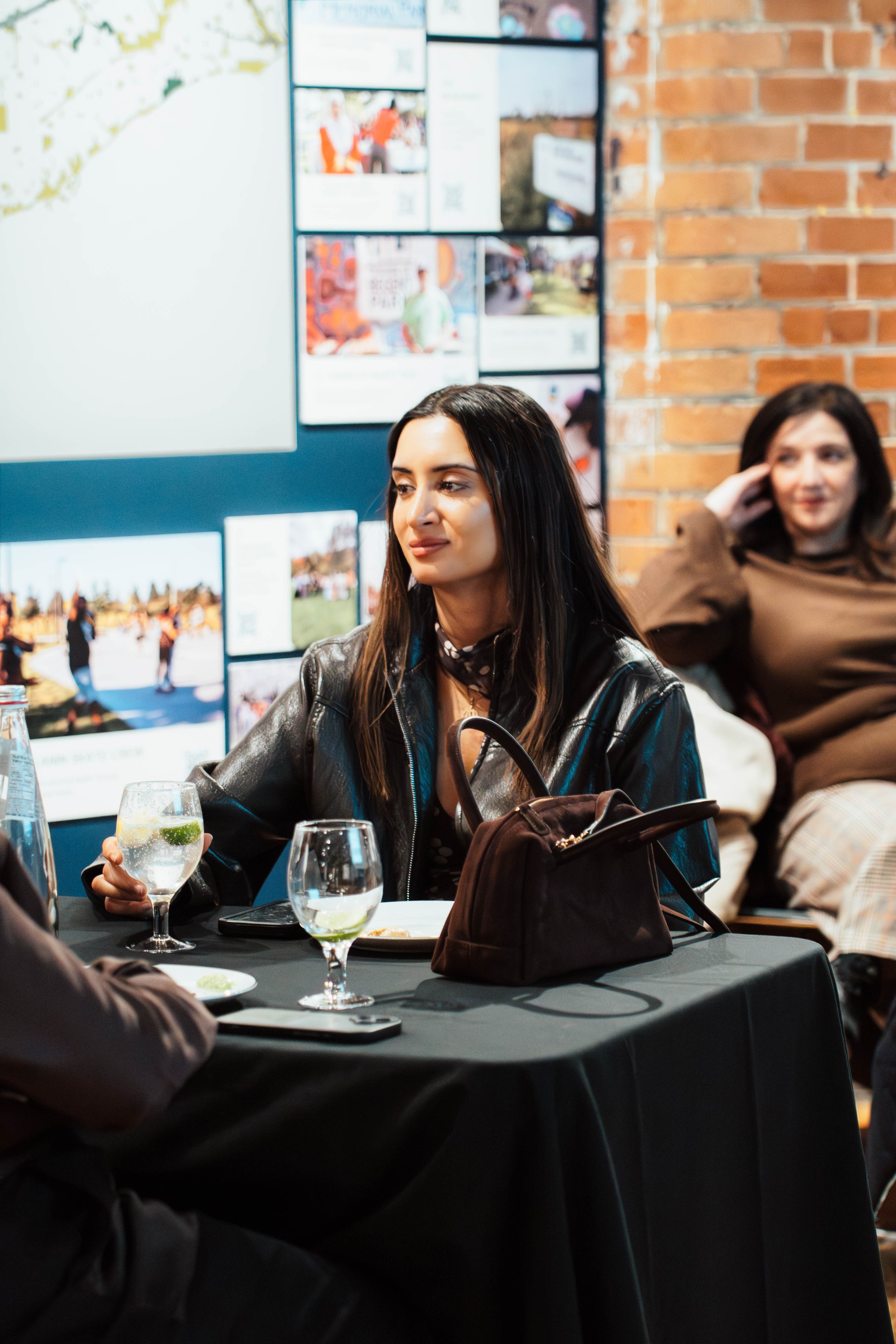 A woman in a leather jacket sitting at a table with drinks and a purse, in a room with a brick wall and a large display board with photos and signs.