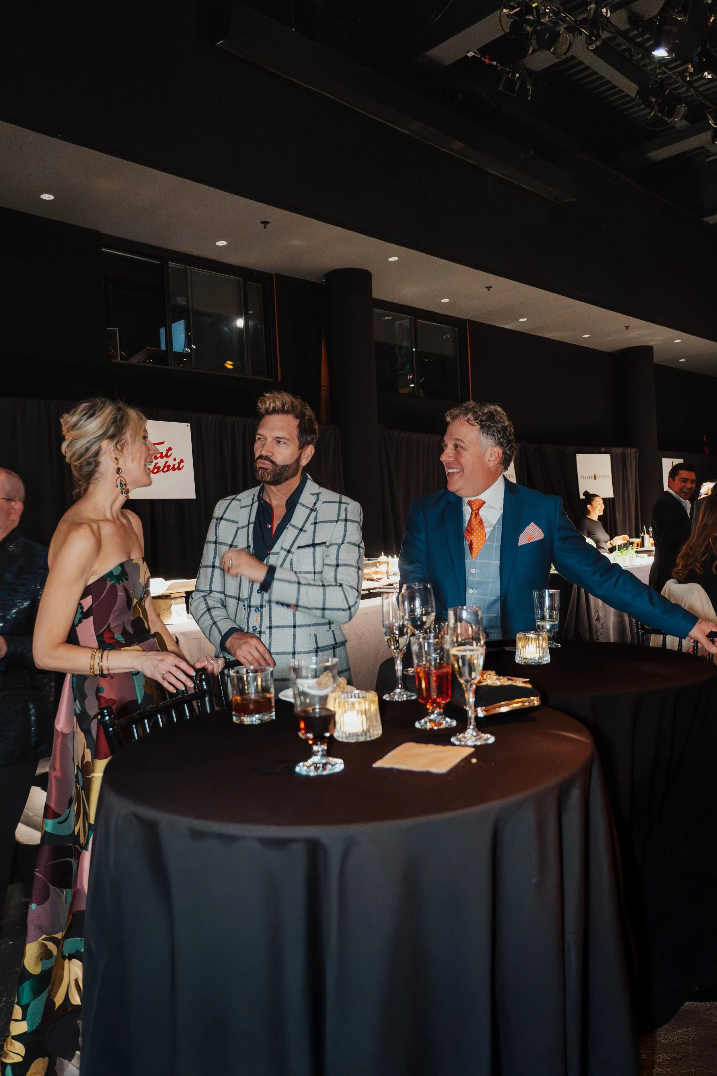 People mingling at a social event or party with drinks on a round table, including a woman in a colorful dress and two men in suits, inside a dimly lit venue.