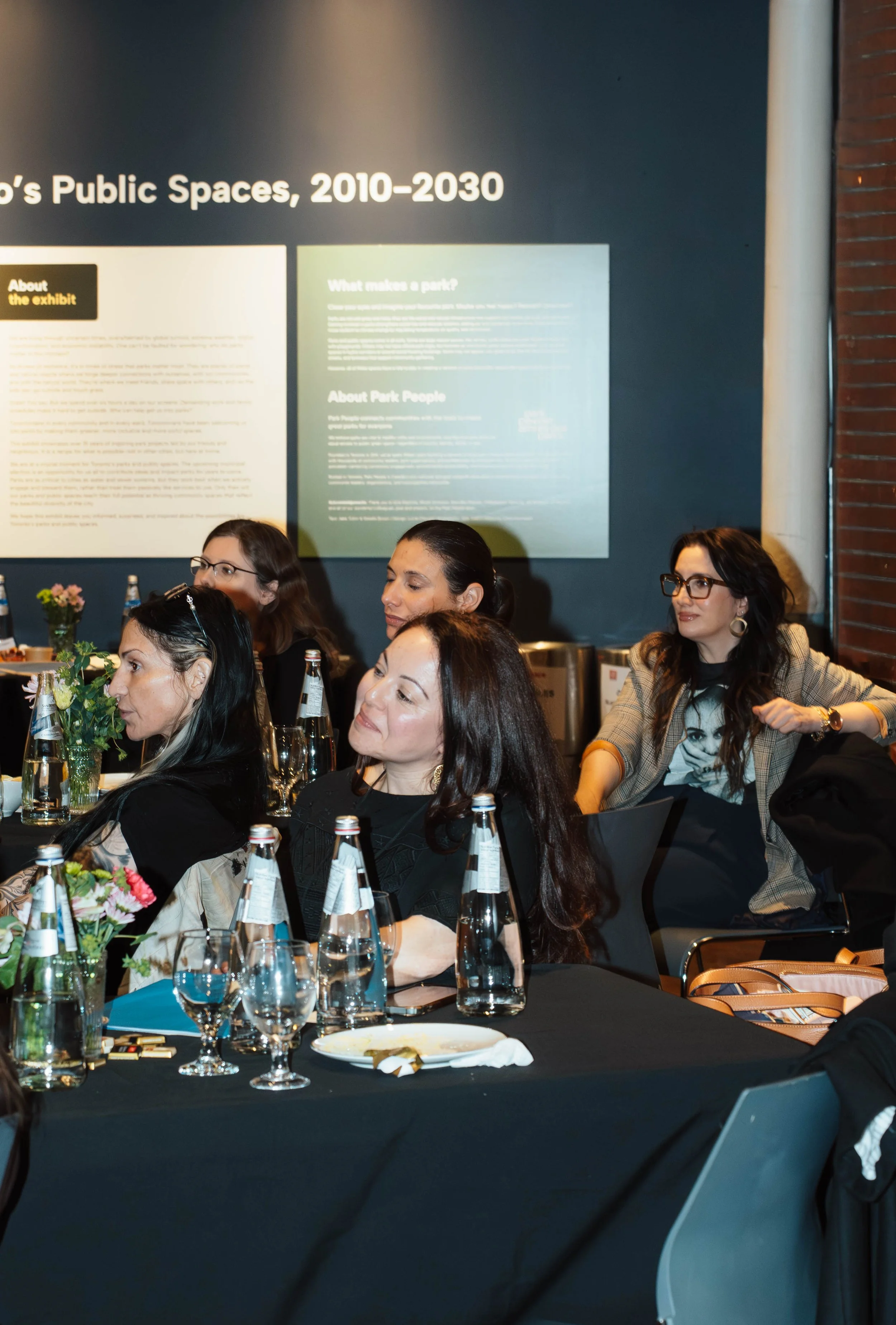 Women seated at a black table during a formal event or meeting, with water bottles, wine glasses, and flower decorations, engaging and listening attentively.