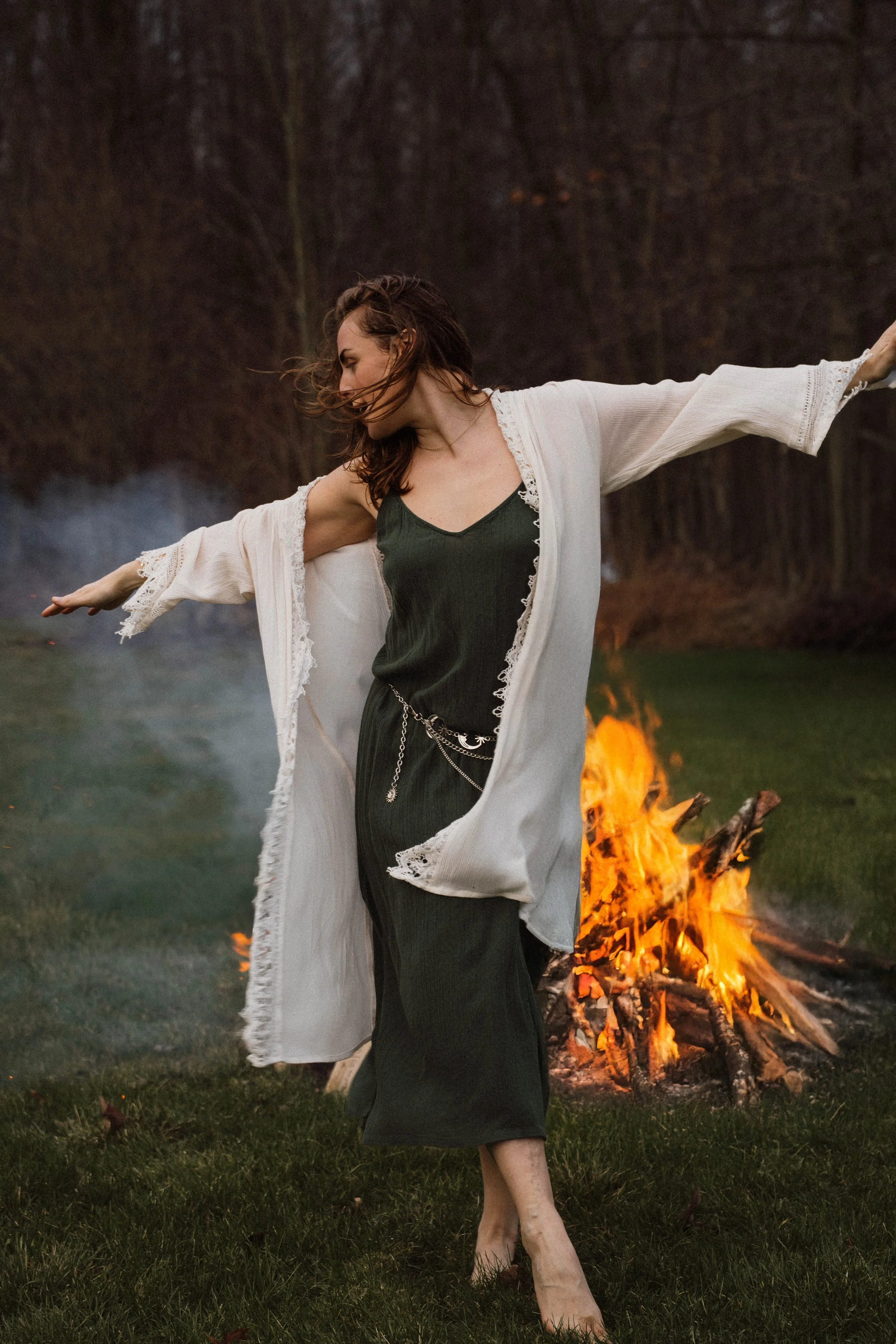 A woman with brown hair dancing barefoot in front of a bonfire on a grassy field, wearing a black dress and a white, loose garment.