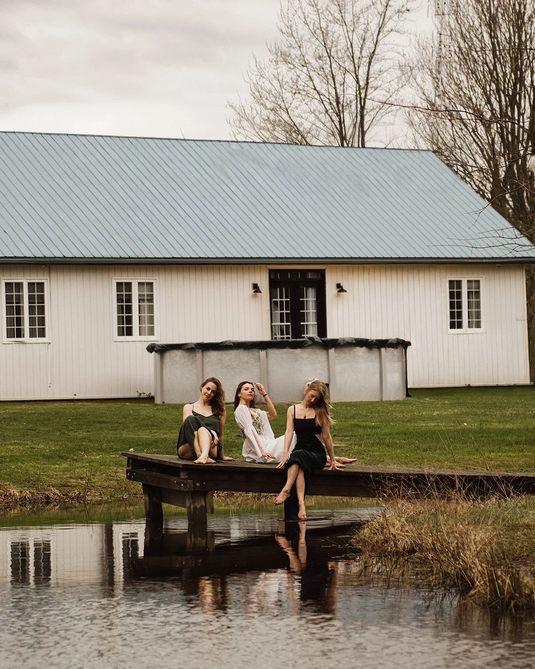 Three women sitting on a wooden dock by a pond with a white barn and leafless trees in the background.