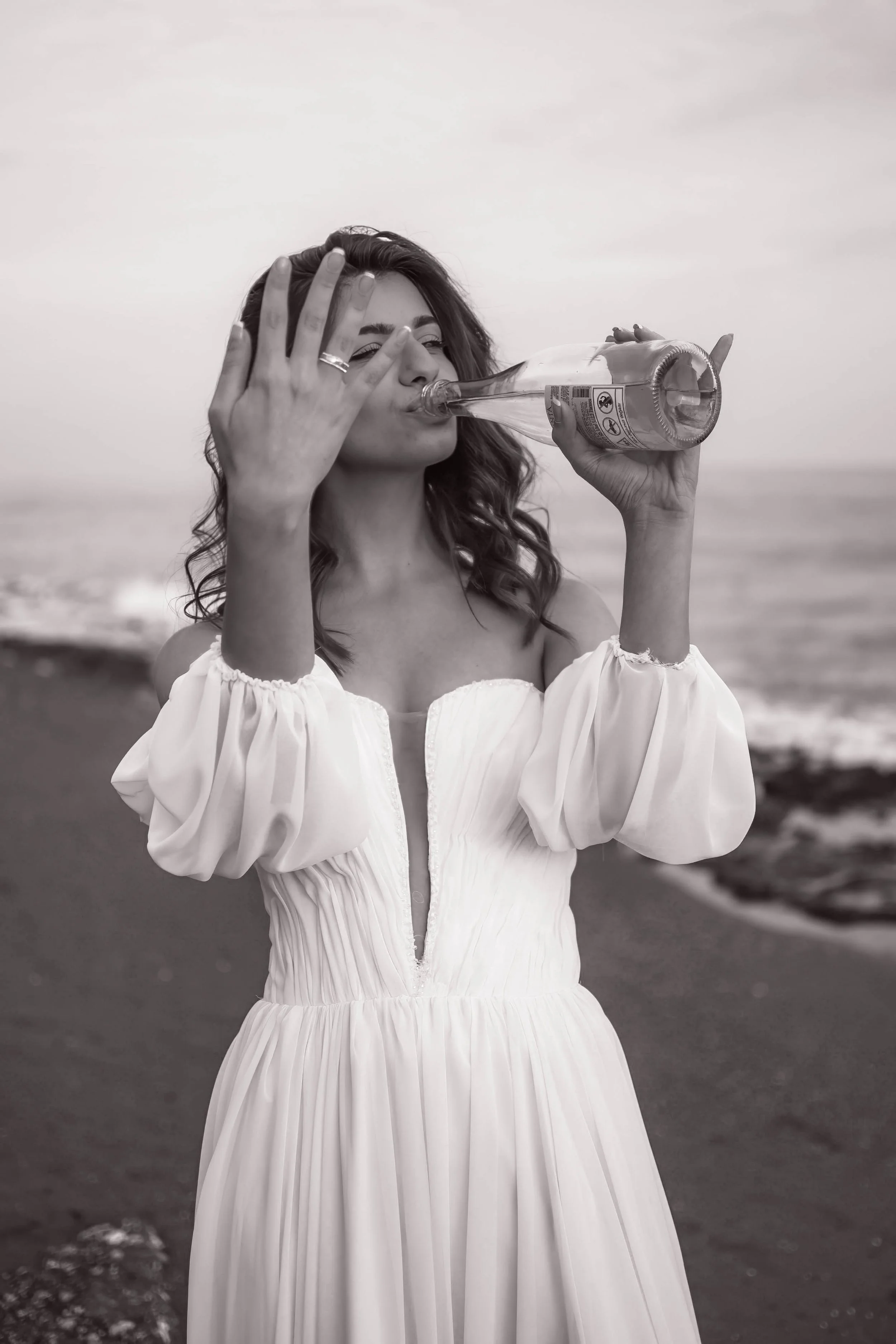 A woman in a white dress drinking from a glass bottle outdoors near the ocean.