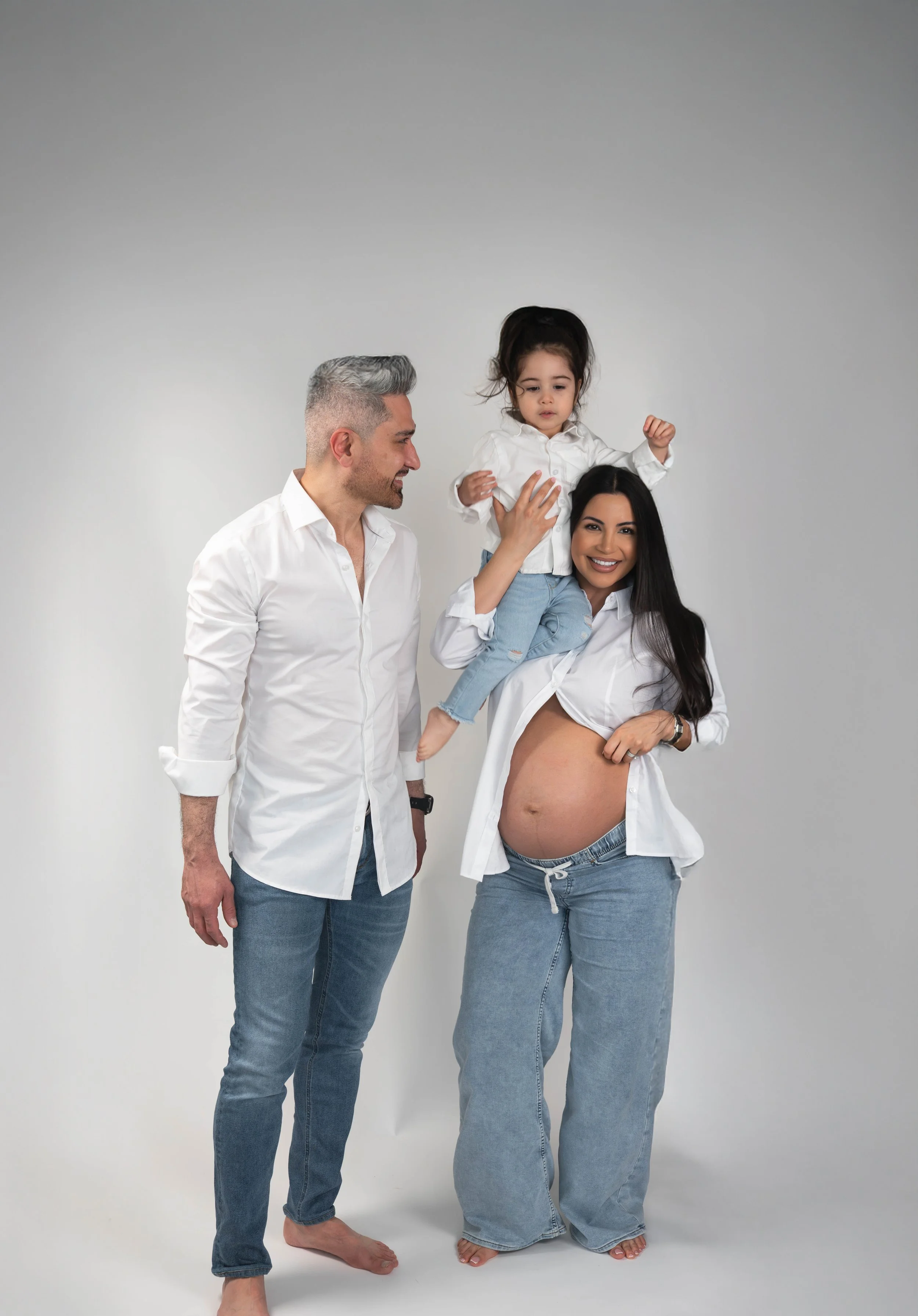 A family of three with a pregnant woman, a man, and a young girl, standing against a plain gray wall. The woman holds her pregnant belly, and the man and girl are looking at each other and smiling.