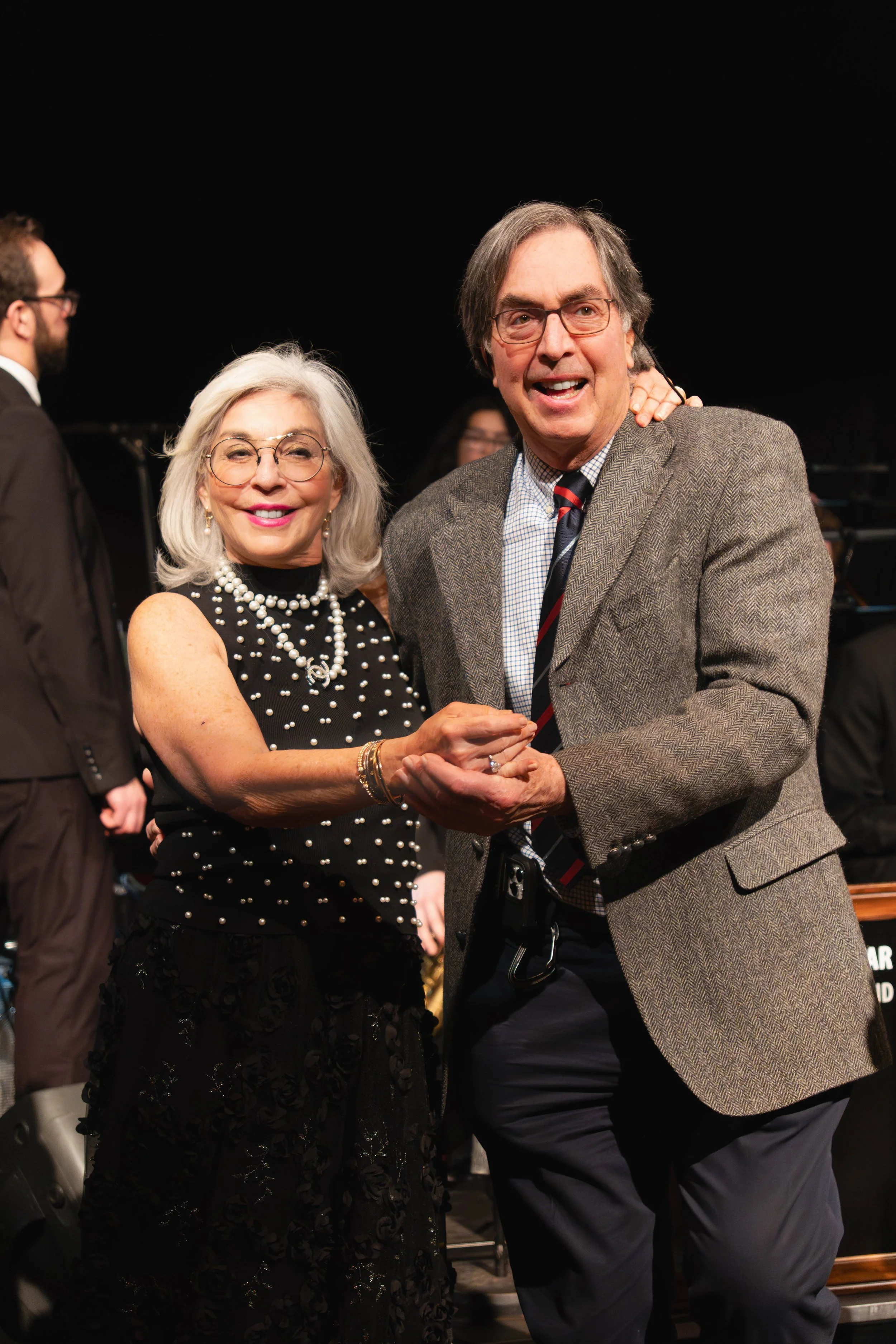 An elderly woman and man dancing together at a formal event, smiling and holding hands. The woman has shoulder-length white hair and glasses, wearing a black dress with pearl embellishments and jewelry. The man has gray hair, wearing glasses, a gray 