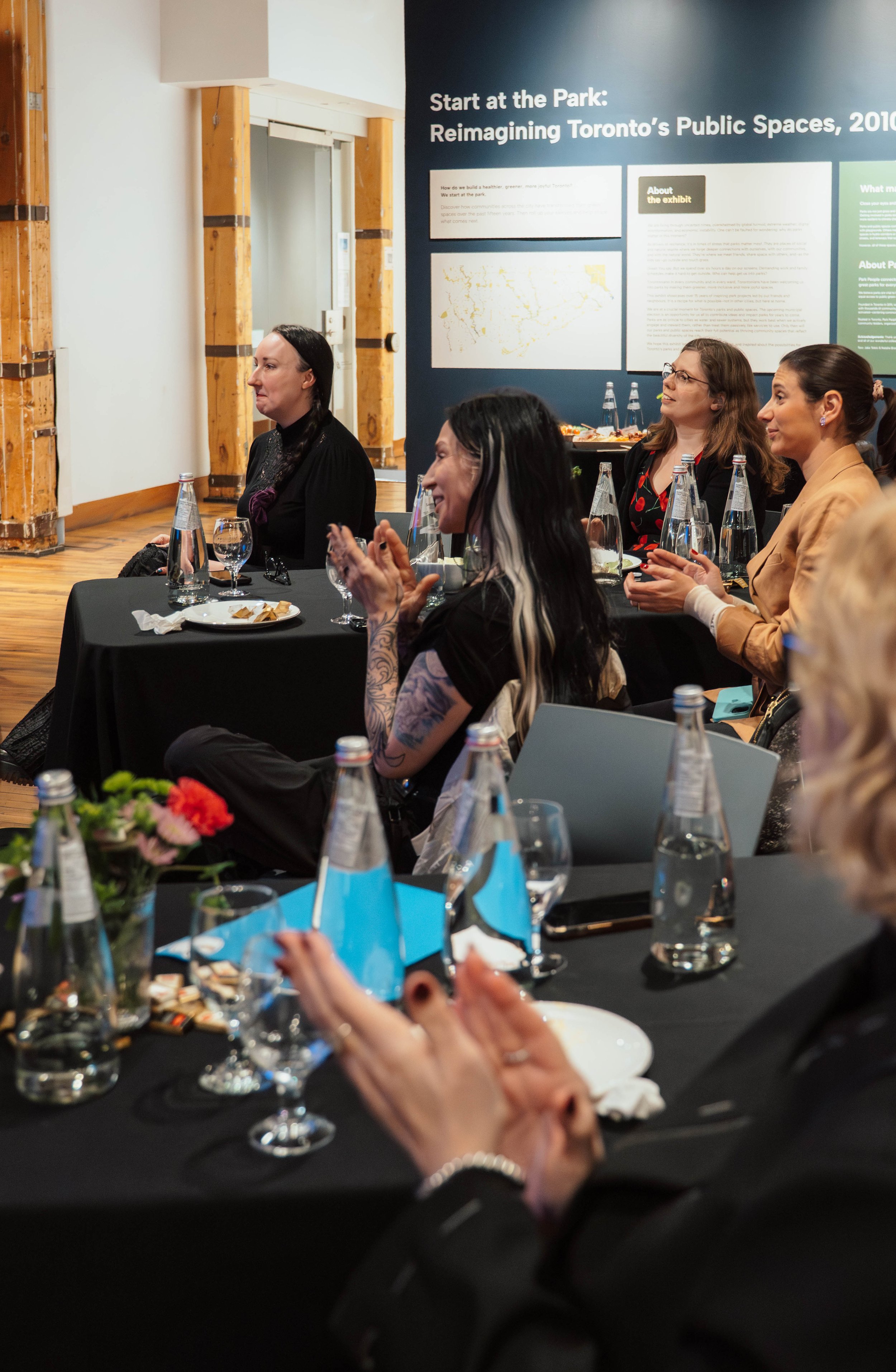 Women sitting at a conference table during a presentation, with water bottles, glasses, and plates on the table, while a wall with informational posters and a map can be seen in the background.