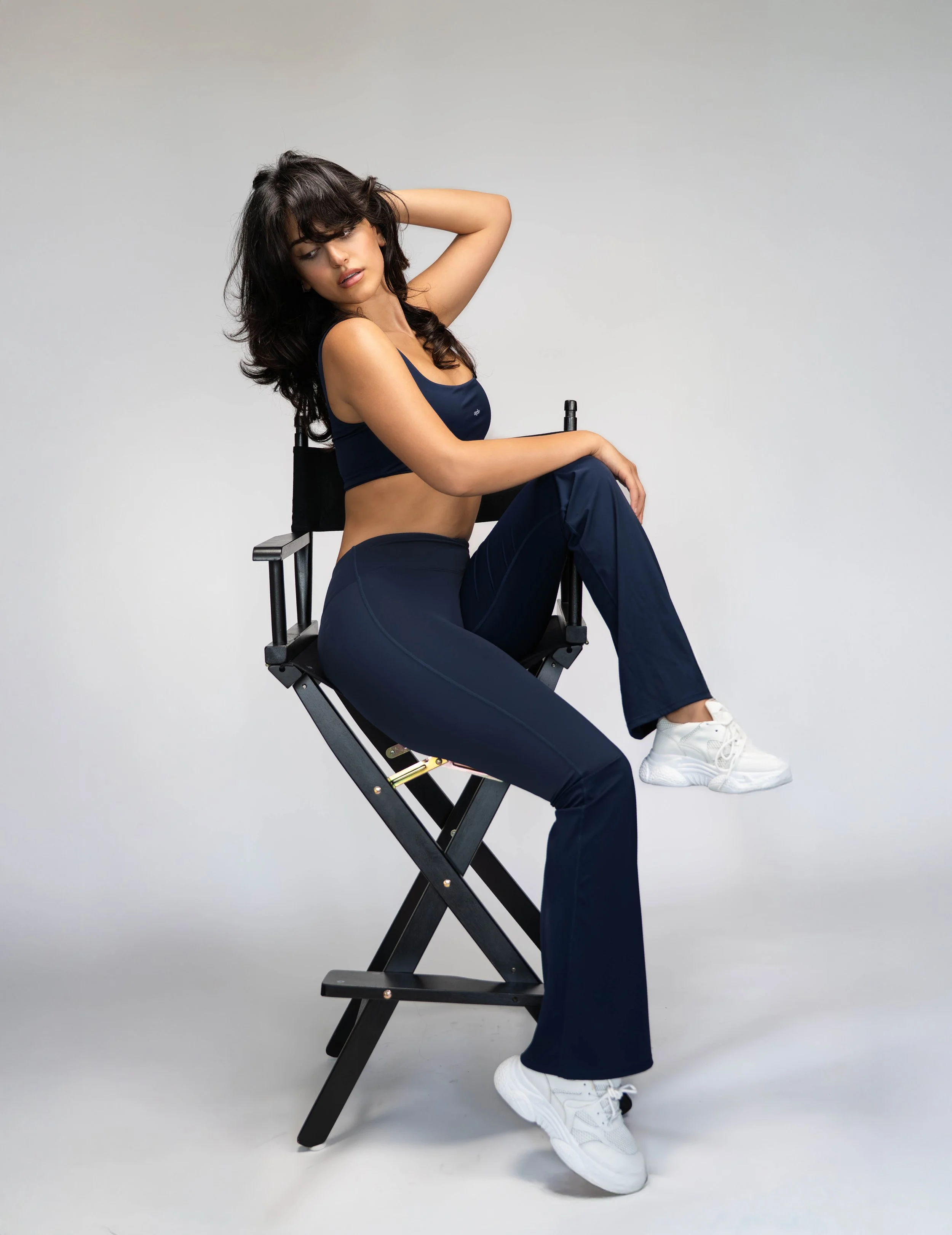 A woman sitting on a director's chair, wearing a black sports bra, matching black leggings, and white sneakers, with her left hand touching her head and her right arm resting on her knee, against a plain light gray background.