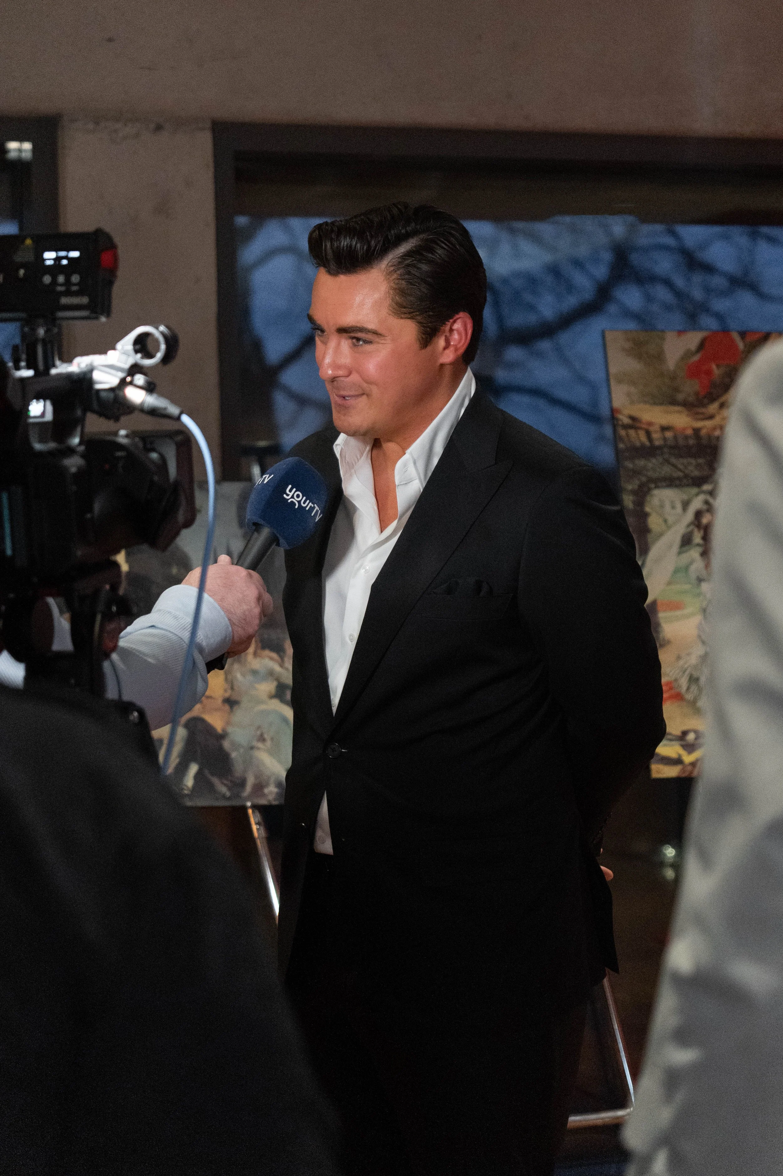 A man in a black suit and white shirt being interviewed by a news crew. The news crew is holding a microphone labeled "YourTV". The background shows a blurry outdoor scene with trees and artwork.