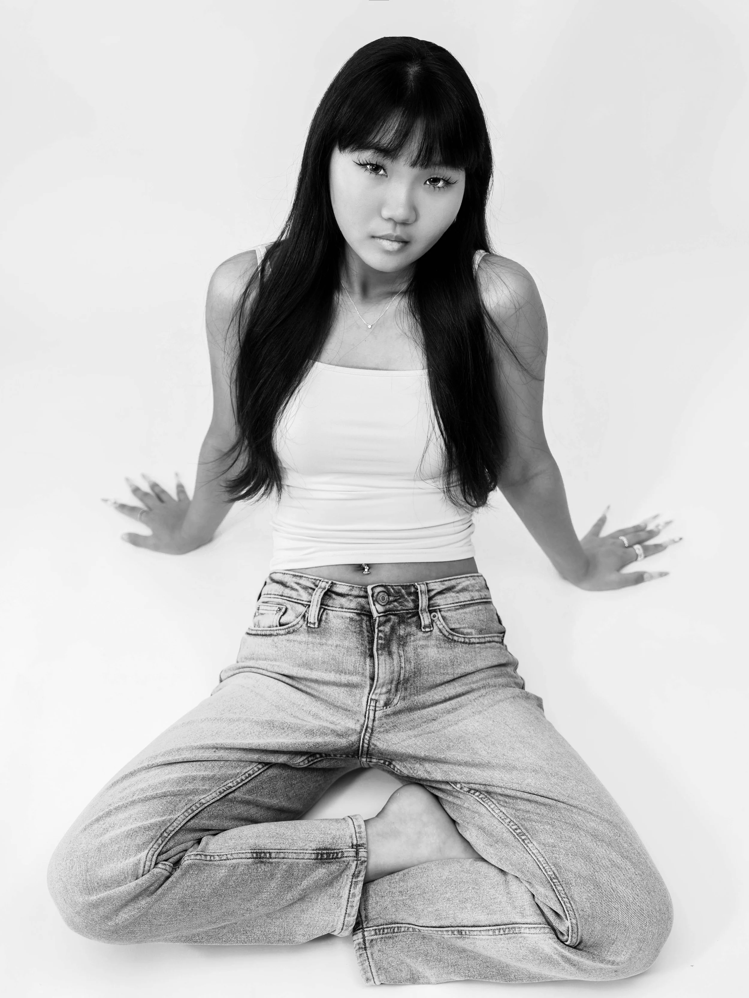 Black and white photo of a woman with long dark hair, sitting with legs crossed and hands on the ground, wearing a white tank top and jeans, looking directly at the camera.