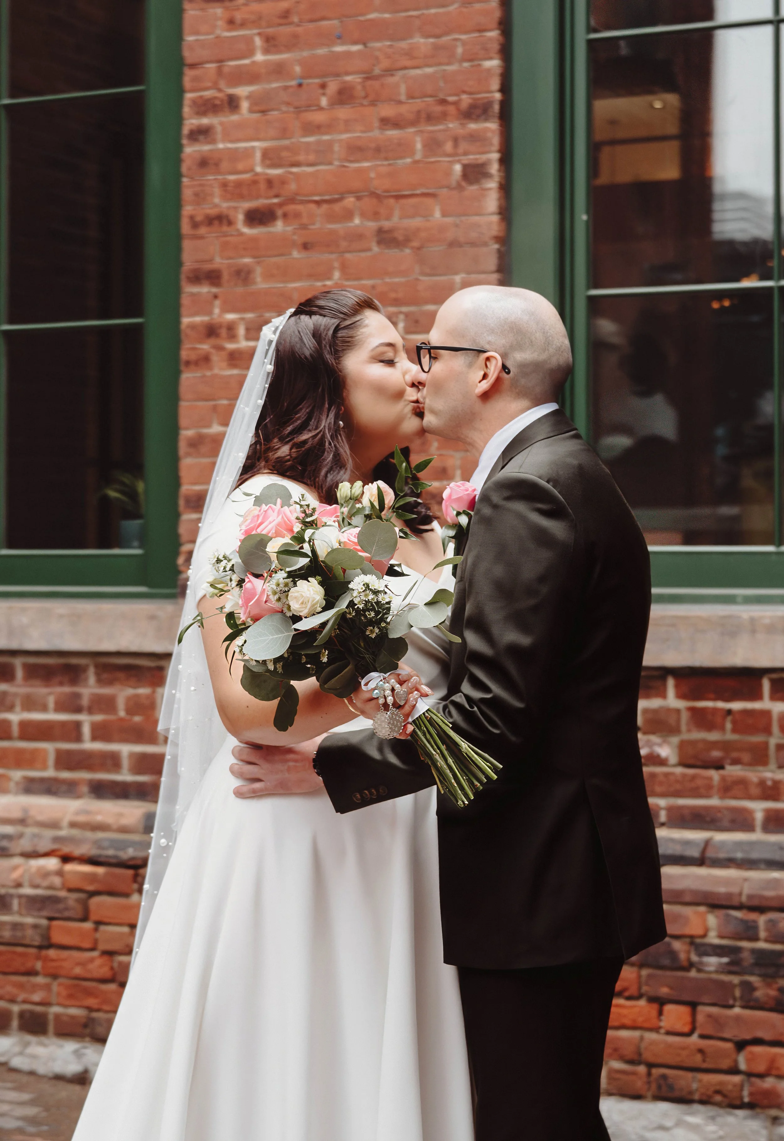 A newlywed couple sharing a kiss outdoors in front of a brick building with green window frames. The bride is holding a bouquet of pink and white flowers, and the groom is dressed in a black suit with glasses.